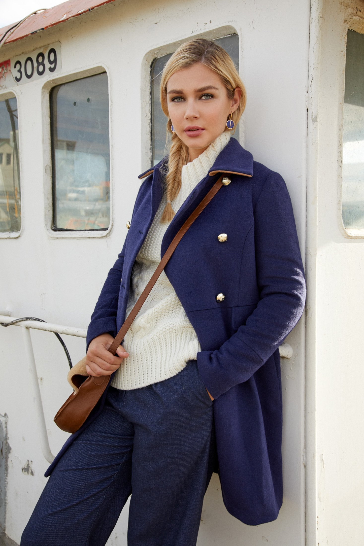 A woman with blonde hair in a braid, wearing a navy blue coat, cream sweater, and blue pants, standing against a white, weathered train or tram with the number 3089, holding a brown purse.
