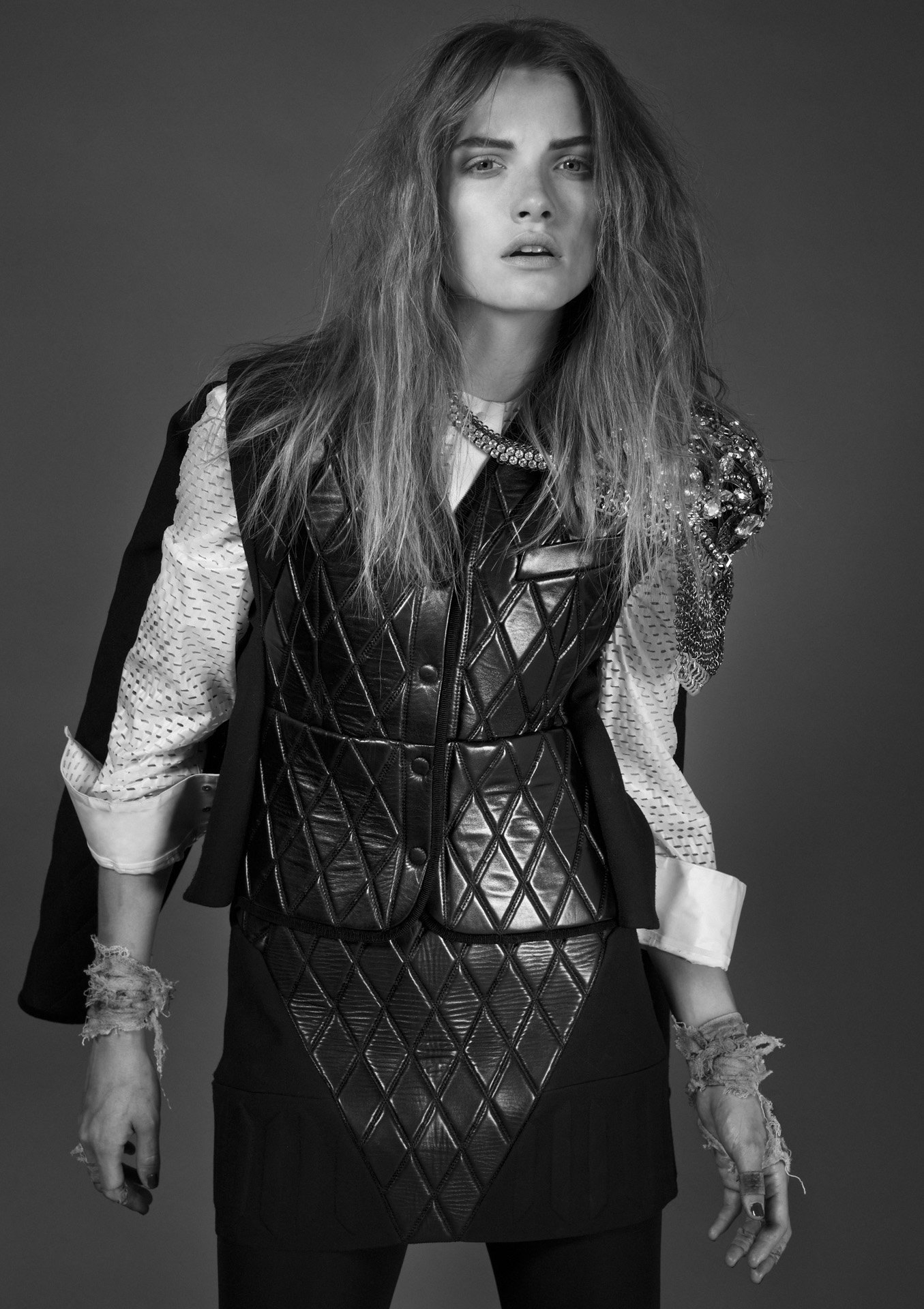 Black and white photo of a woman with long, tousled hair, wearing a quilted leather vest with jeweled embellishments, layered over a patterned shirt with frayed sleeves, standing against a plain background.