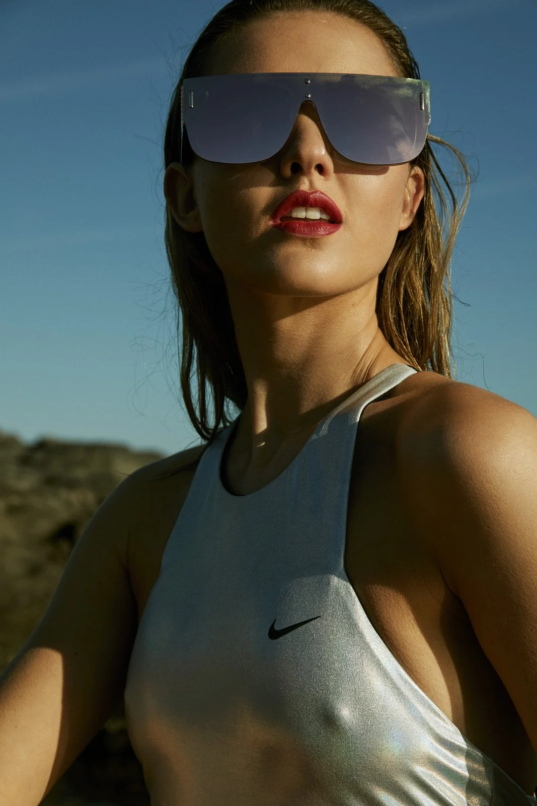A woman wearing reflective sunglasses, red lipstick, a white Nike tank top, with a rocky landscape and clear blue sky in the background.