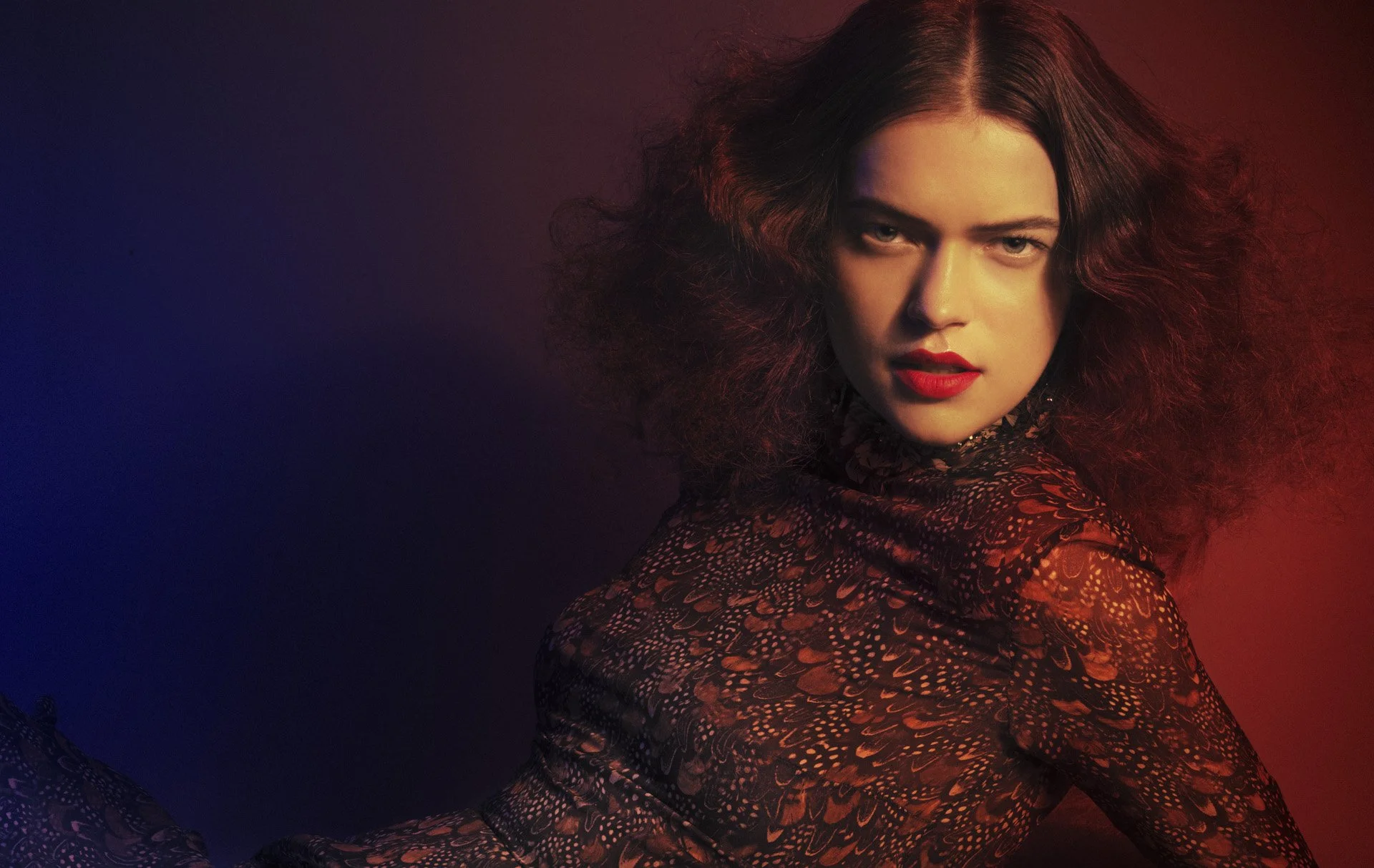 A woman with voluminous hair, red lipstick, and a patterned dress looking fiercely at the camera against a gradient background of blue, purple, and red.