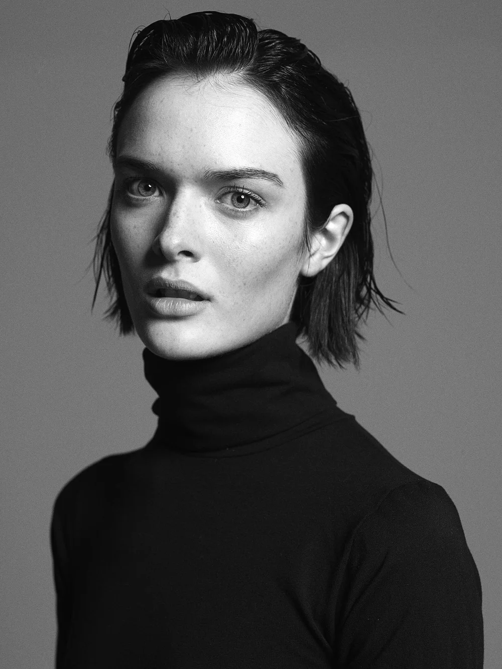 Black and white portrait of a woman with wet, slicked-back hair, wearing a black turtleneck, looking directly at the camera with a neutral expression.