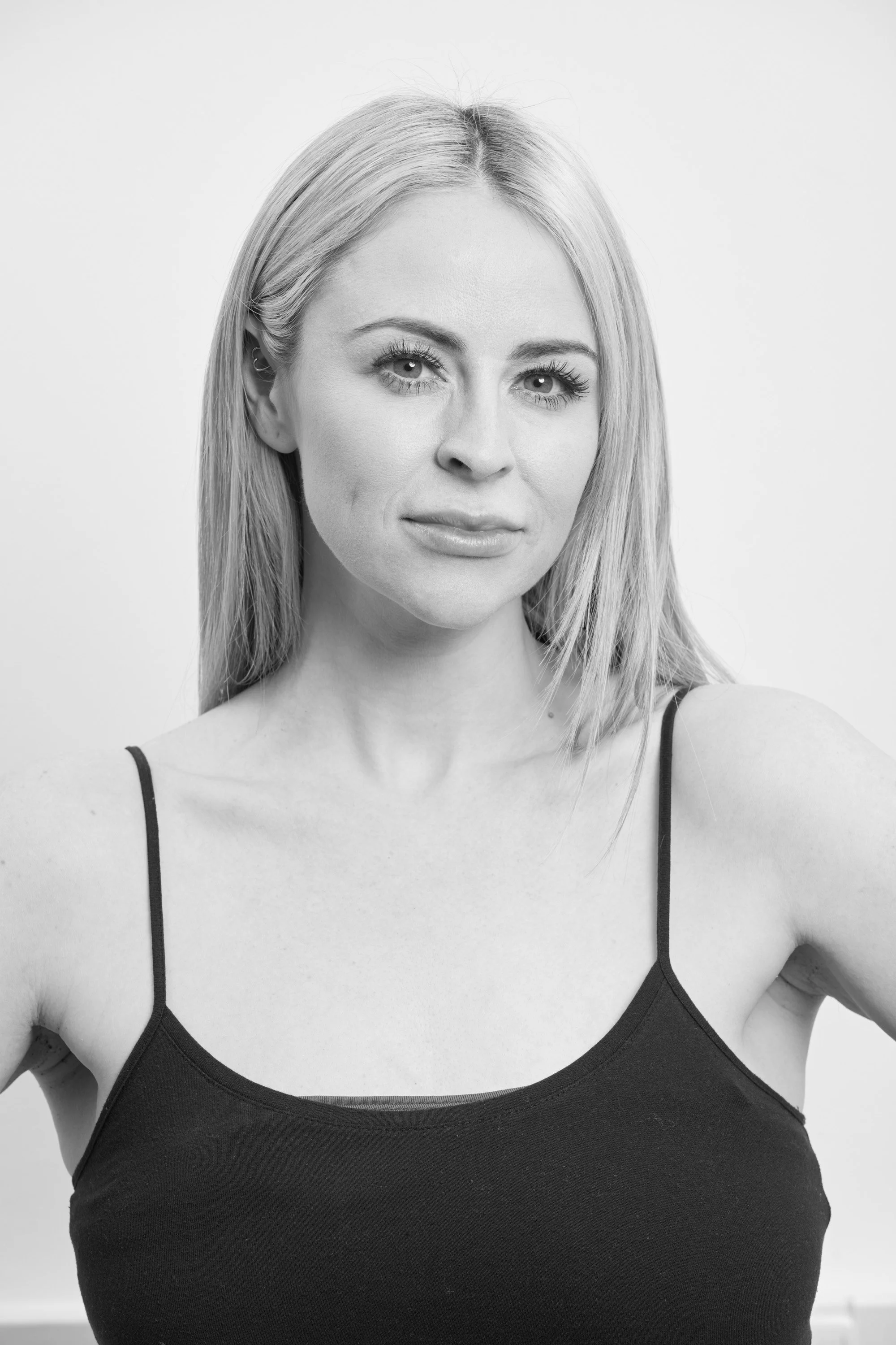Black and white portrait of a woman with blonde hair wearing a dark spaghetti strap top, smiling slightly, with a neutral background.