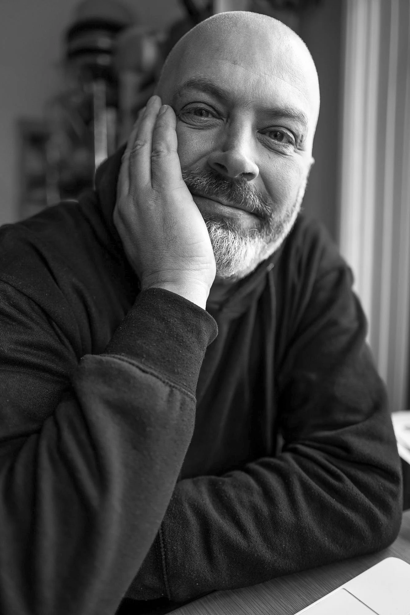 A black and white photo of a bald man with a beard, smiling, resting his face on his hand, sitting at a table with a laptop.