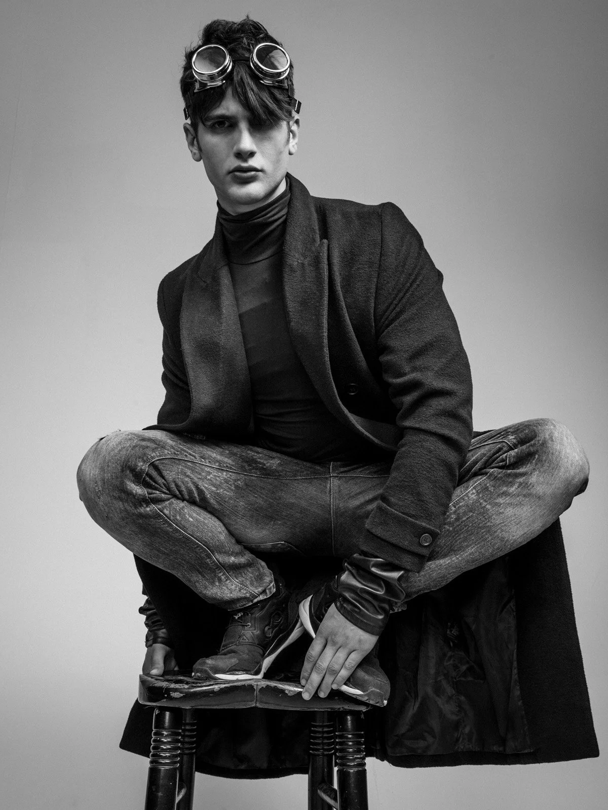 Black and white photo of a young man sitting cross-legged on a stool, wearing a dark blazer, a turtleneck, and jeans. He has glasses on his head and is looking directly at the camera with a serious expression.