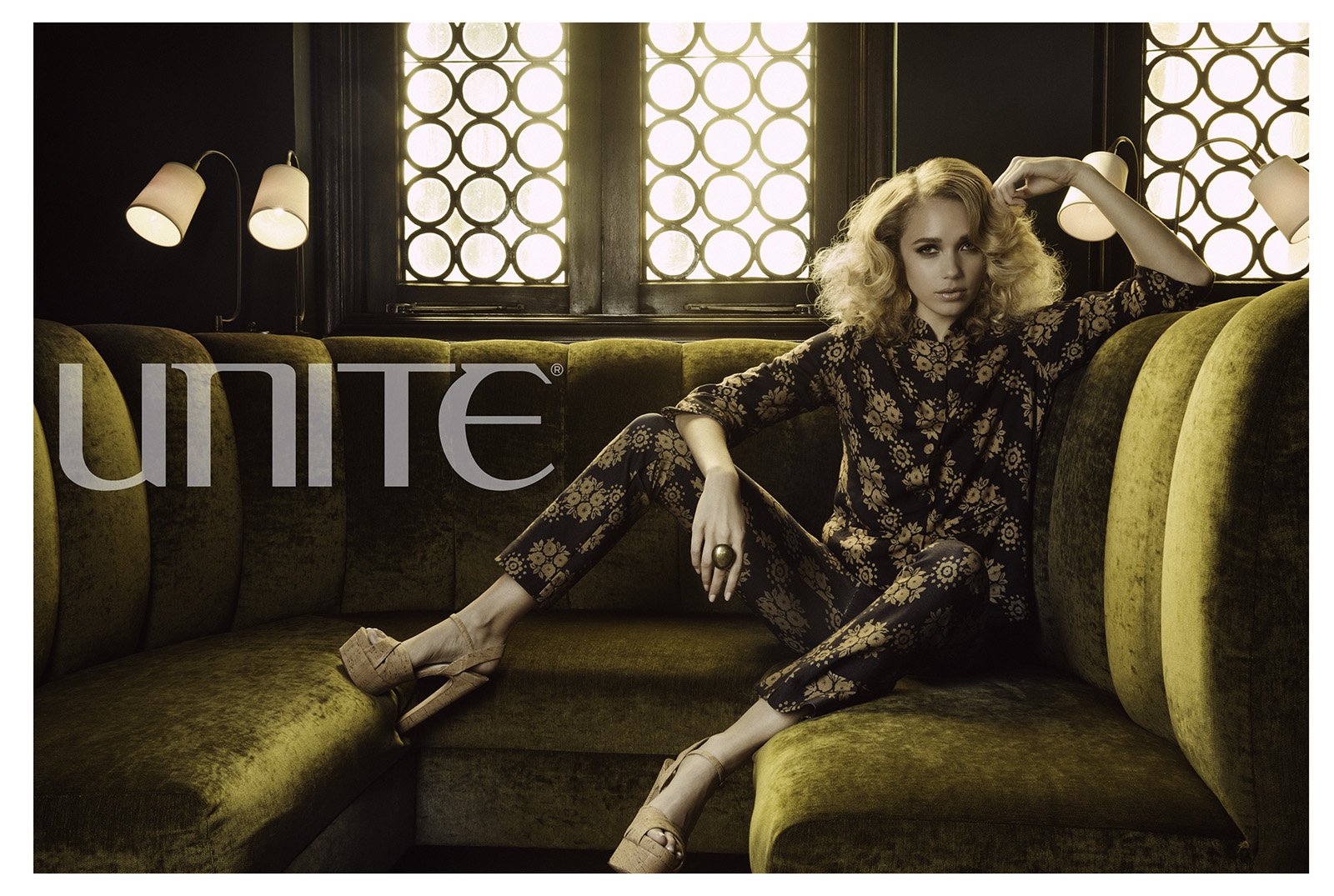 A woman with curly blonde hair sitting on a green velvet sofa in a dimly lit room with stained glass windows and wall lamps. She is wearing a black floral suit, beige high heels, and a large ring, posing with one arm raised and a confident expression