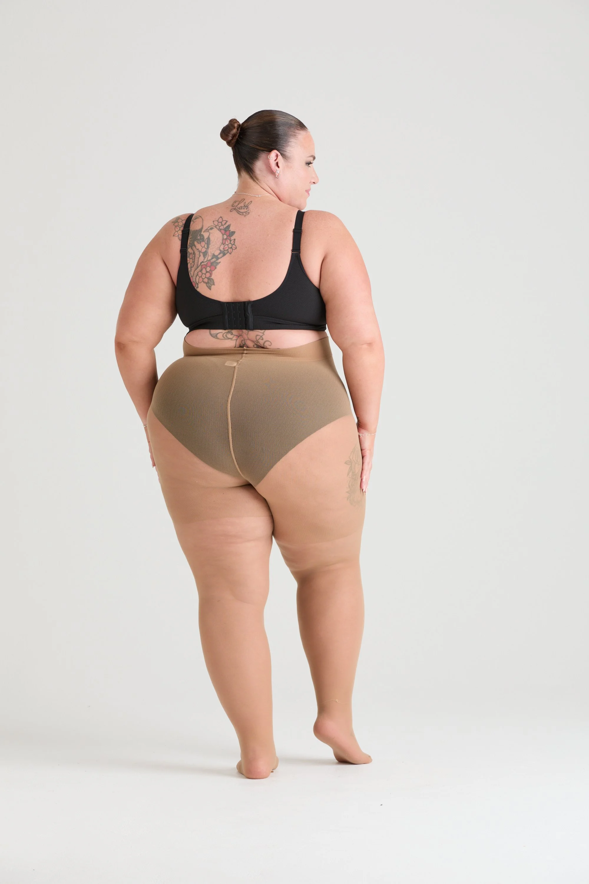 A woman with dark hair in a bun, tattoos on her back and arm, wearing black bra and tan shapewear, standing on her toes against a plain white background.