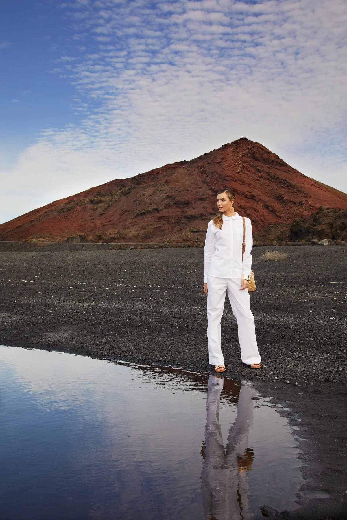 A woman in a white outfit standing on dark volcanic sand near a reflective pool, with a reddish mountain and cloudy sky in the background.