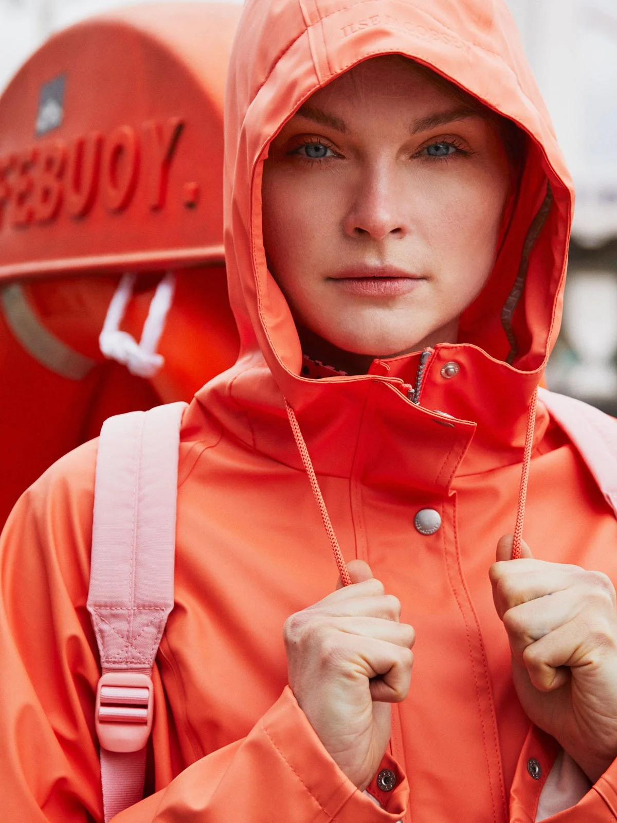 A woman dressed in a bright orange rain jacket and matching hood, holding the straps of a pink backpack, with a life-preserving buoy in the background labeled 'SIBOY'.