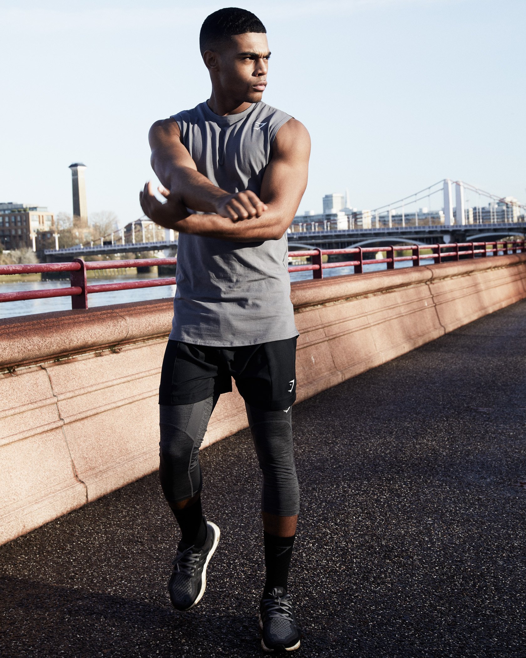 Young man stretching outdoors on a bridge during daytime, wearing athletic clothing and running shoes.