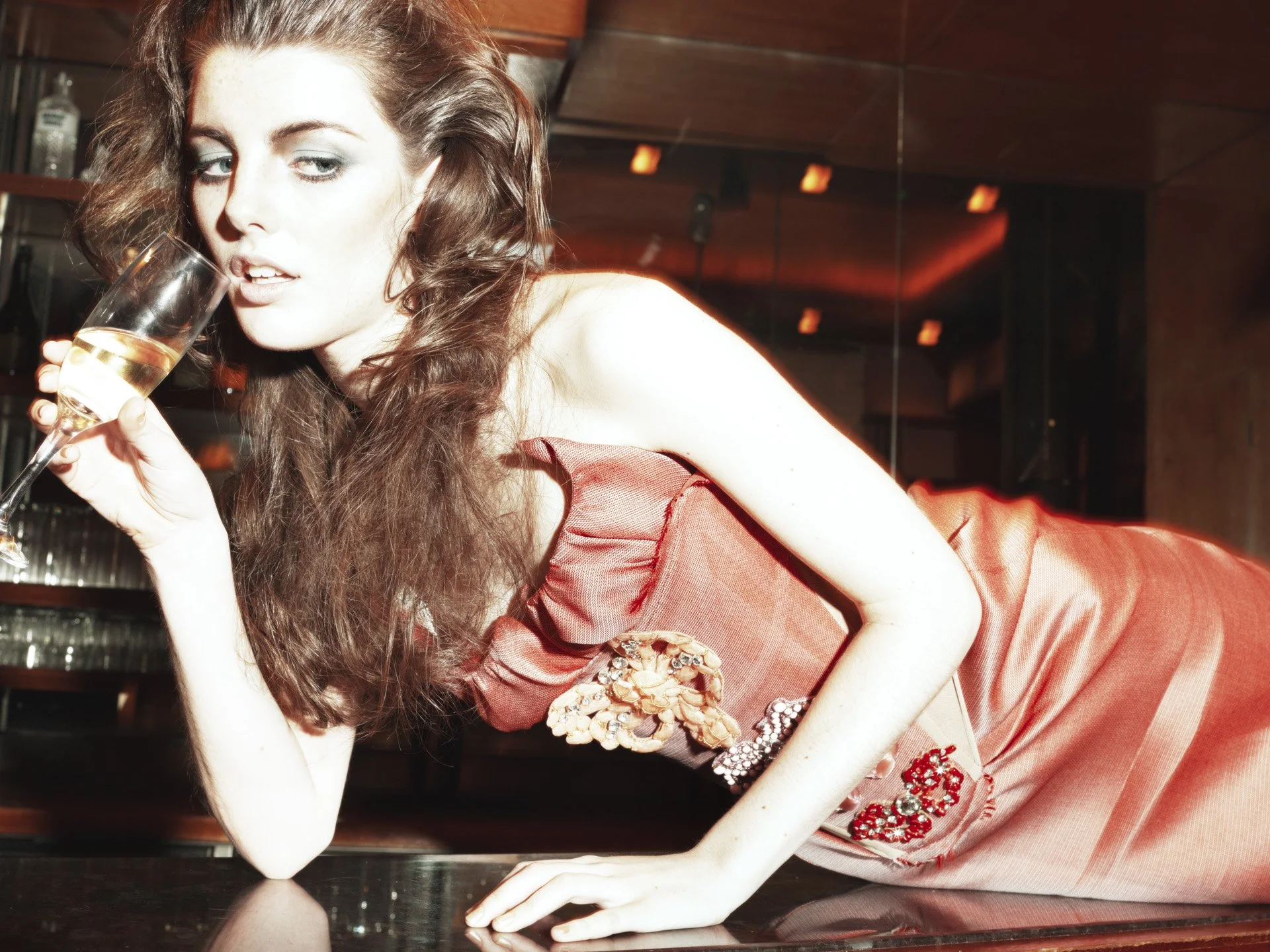 A woman with long, wavy brown hair and fair skin, wearing a reddish-orange dress with floral embellishments, is lying on a reflective surface in a dimly lit room with warm lighting. She is holding a glass of sparkling wine near her lips and looking a