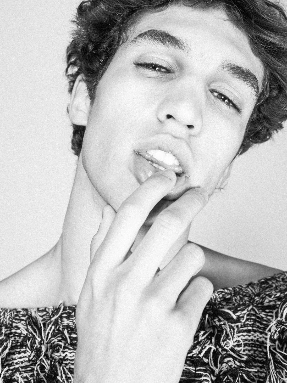 Close-up black and white photograph of a young woman with short, curly hair. She is touching her lips with her fingers, slightly parted, and gazing into the camera with a sensual expression. She is wearing a patterned top.