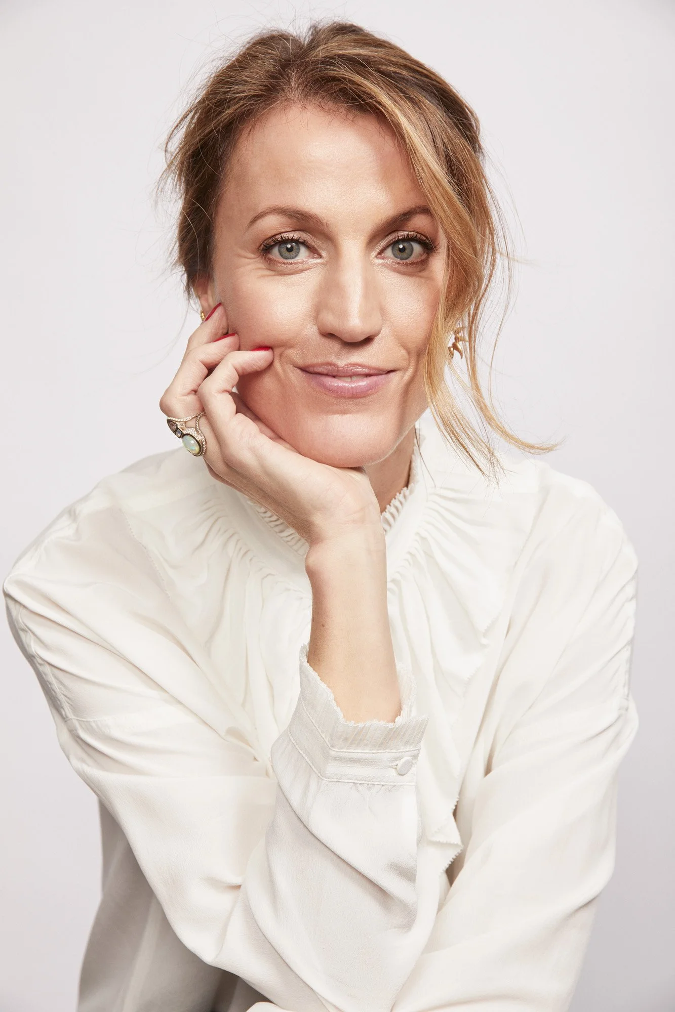 A woman with blonde hair and blue eyes, wearing a cream-colored shirt, resting her head on her hand, looking directly at the camera with a subtle smile.