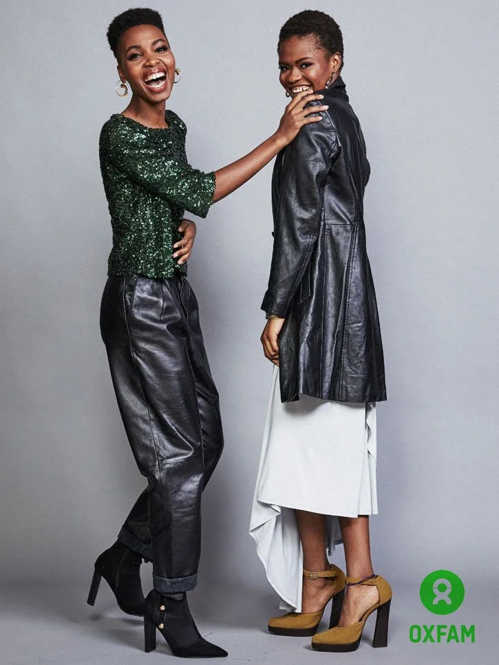 Two women smiling and laughing, one wearing a green sequined top and black leather pants, the other in a black leather coat and white skirt, standing against a plain gray background with an Oxfam logo.
