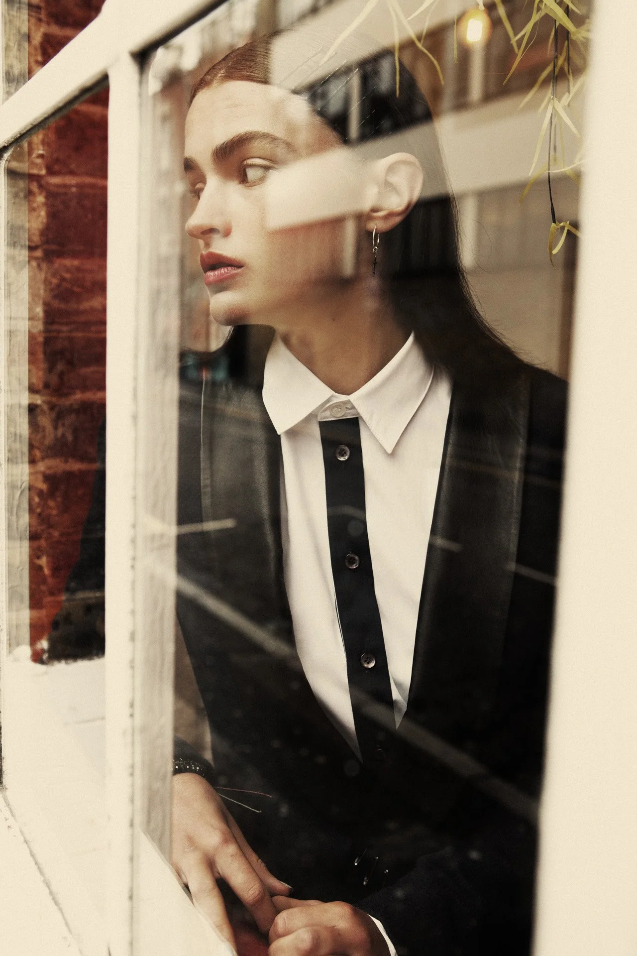 A woman with long dark hair and earrings looking out a window, wearing a white shirt with a black blazer, viewed through the glass window.