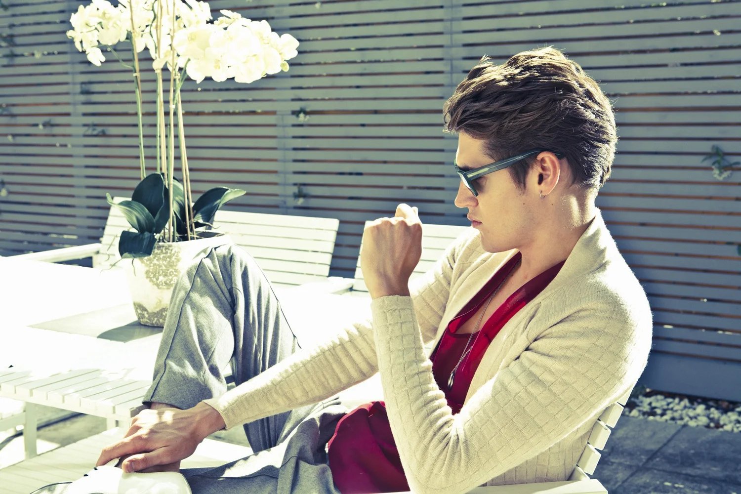 A young man wearing sunglasses, a beige blazer, red shirt, and gray pants sits on a white outdoor lounge chair on a sunny patio, with a large potted plant with white flowers nearby and a wooden fence in the background.