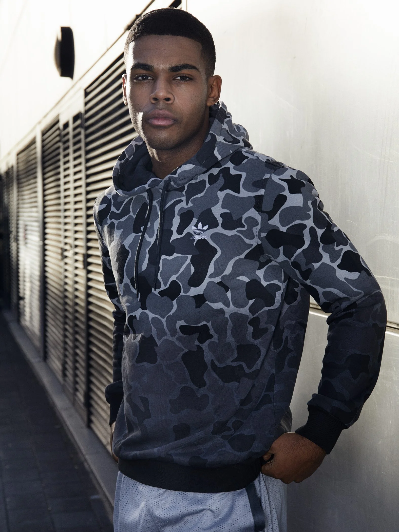 A young man with dark skin and a short haircut wearing a camouflage-patterned Adidas hoodie and gray athletic shorts, standing outdoors against a wall with ventilation panels.