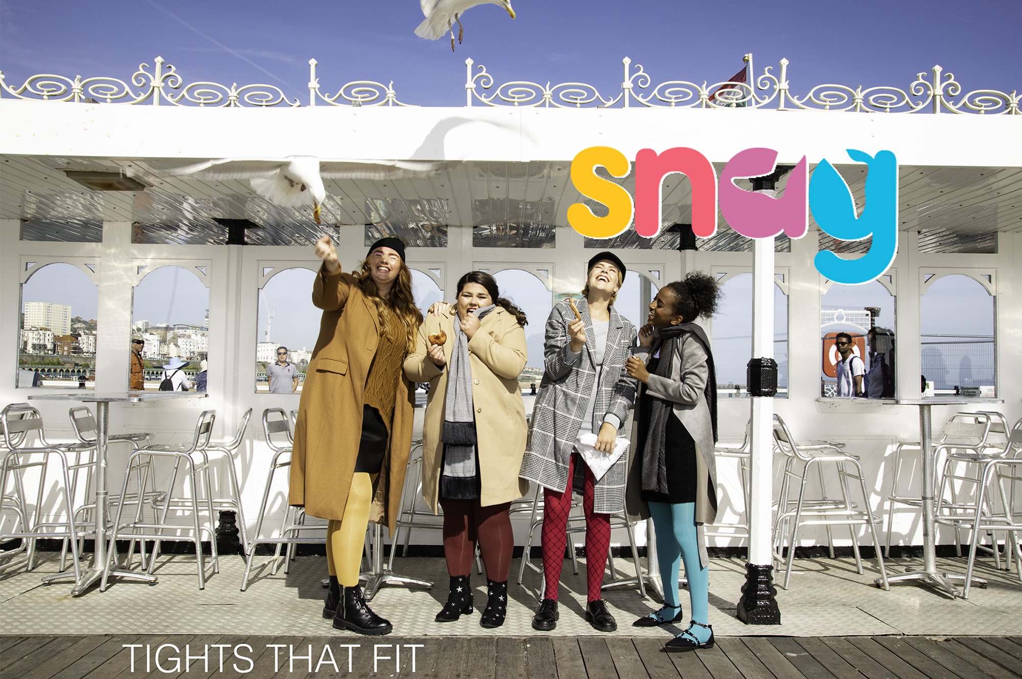 Four women standing together on a boat deck, with a seagull flying overhead. They are smiling and dressed in colorful, fashionable tights and coats. There are chairs and a railing behind them, with a cityscape in the background and a bright blue sky.