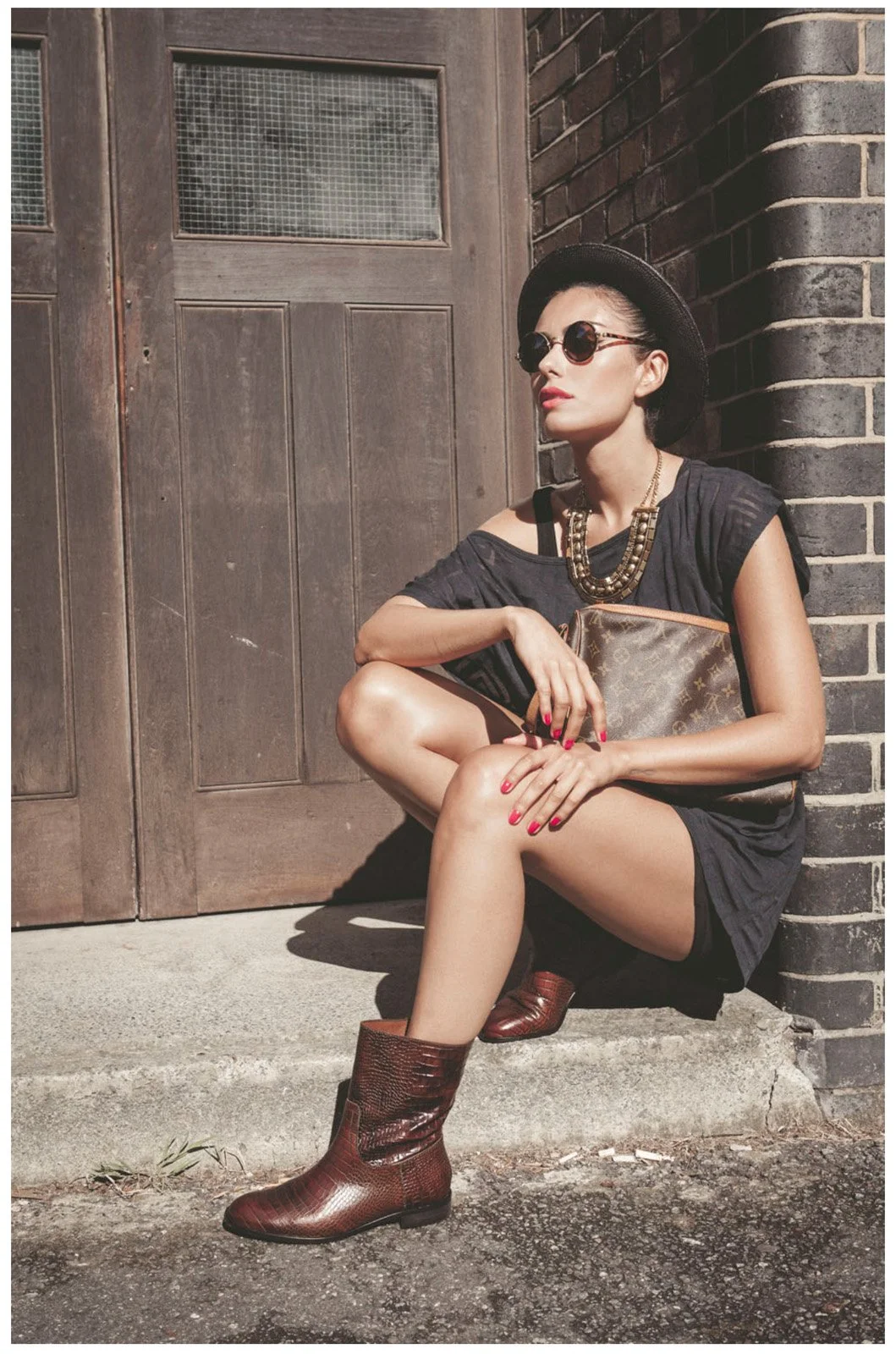 A woman sitting on a sidewalk near a brick building, wearing sunglasses, a black hat, a black dress, brown boots, and holding a designer clutch, with jewelry and red lipstick.