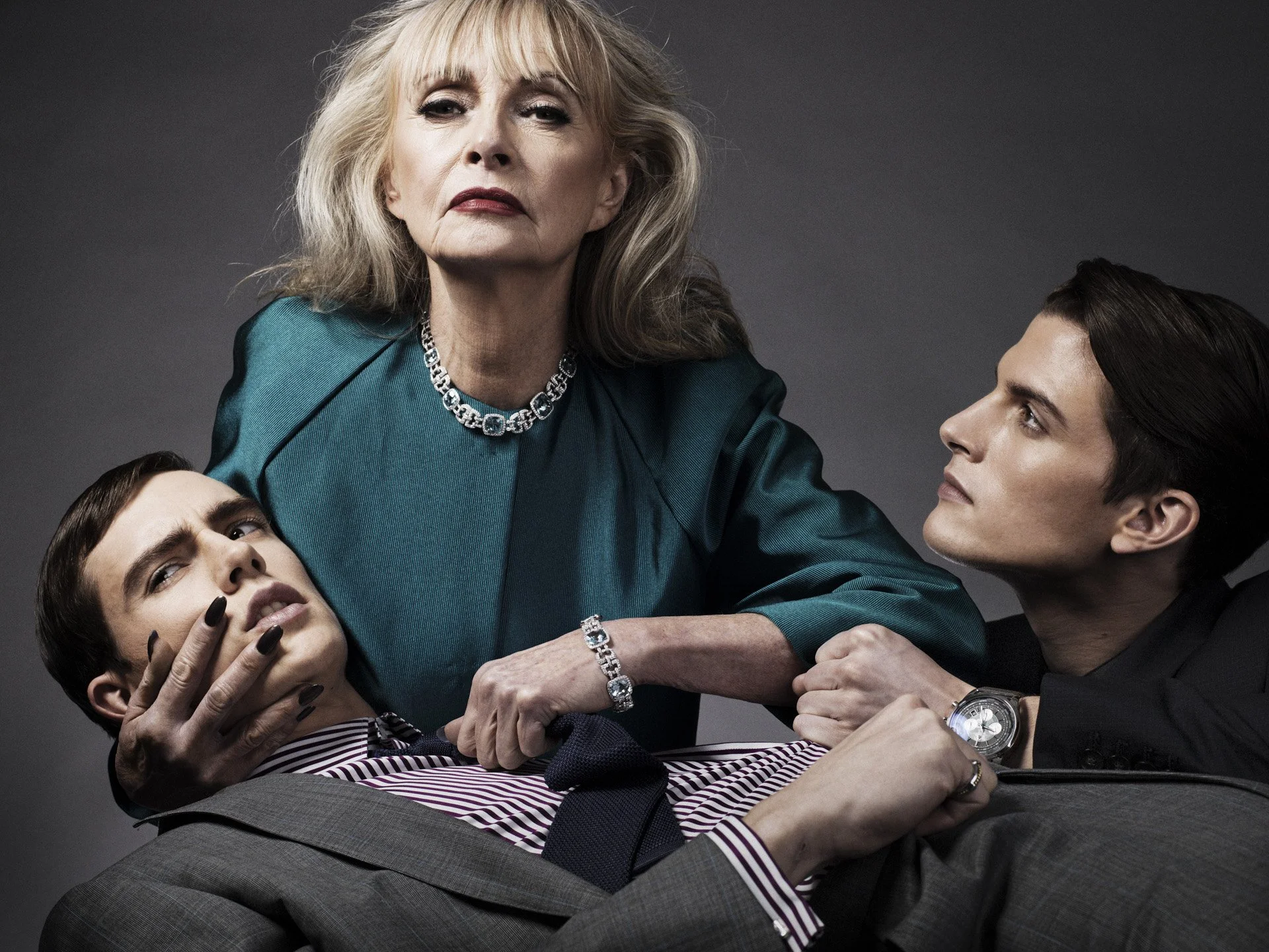 An older woman with blonde hair, wearing a teal dress and diamonds, holds and restrains three young adults. The young woman with dark hair and a striped shirt looks distressed, and the young men with dark hair and watches show concern or surprise.