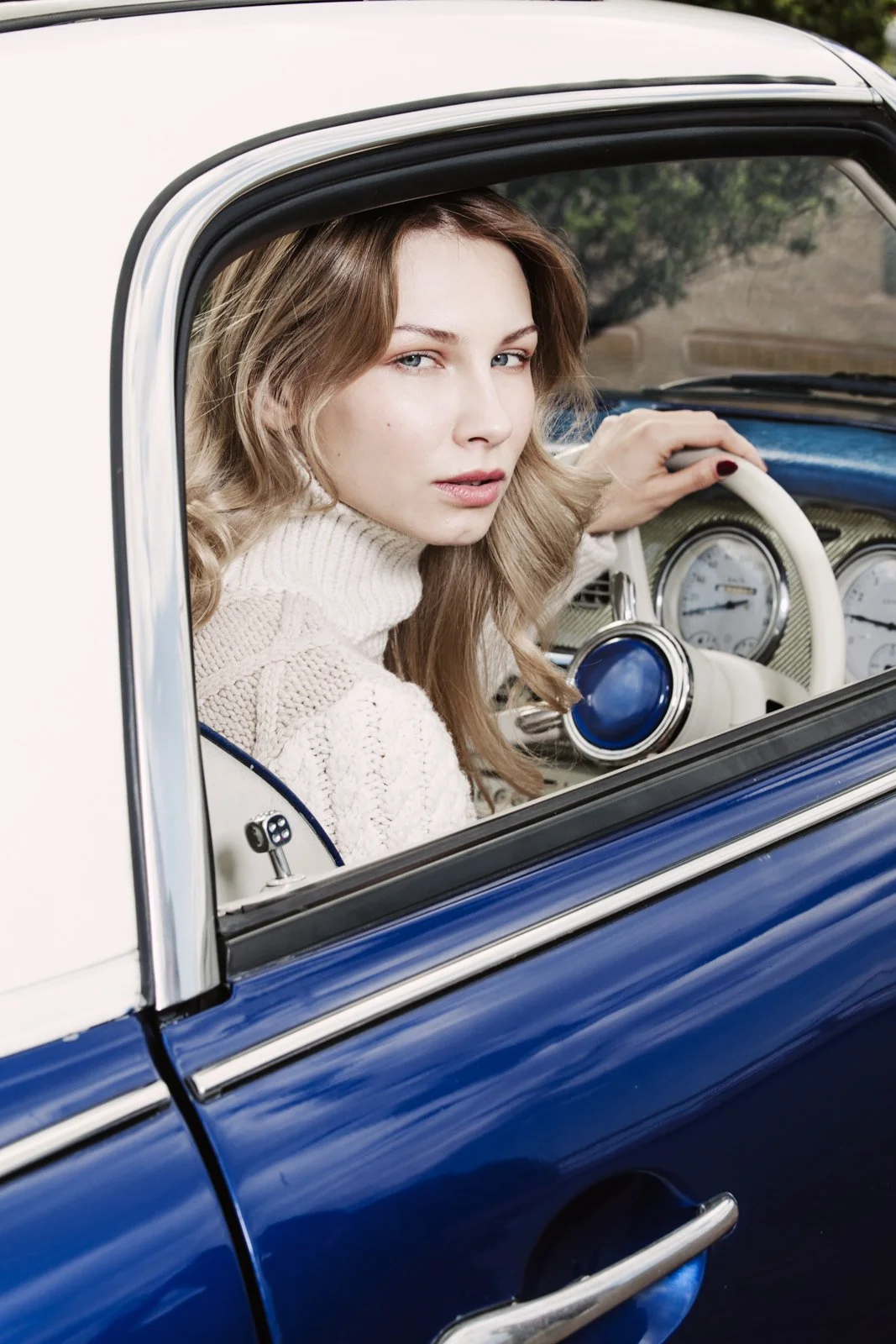 A woman with long wavy blonde hair and a cream-colored knit sweater sitting in the driver’s seat of a vintage blue and white car, looking out the window with a slight smile.