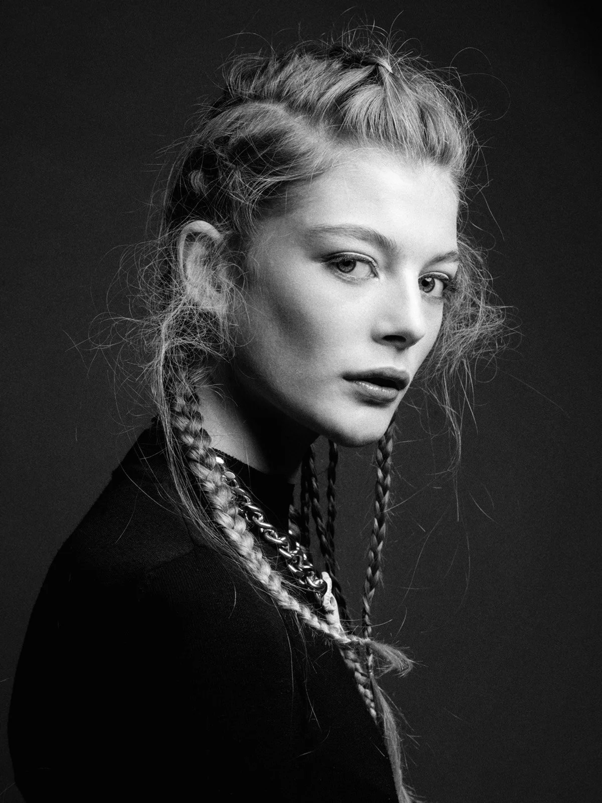 Black and white portrait of a young woman with textured hair and braids, wearing a chain necklace, against a dark background.