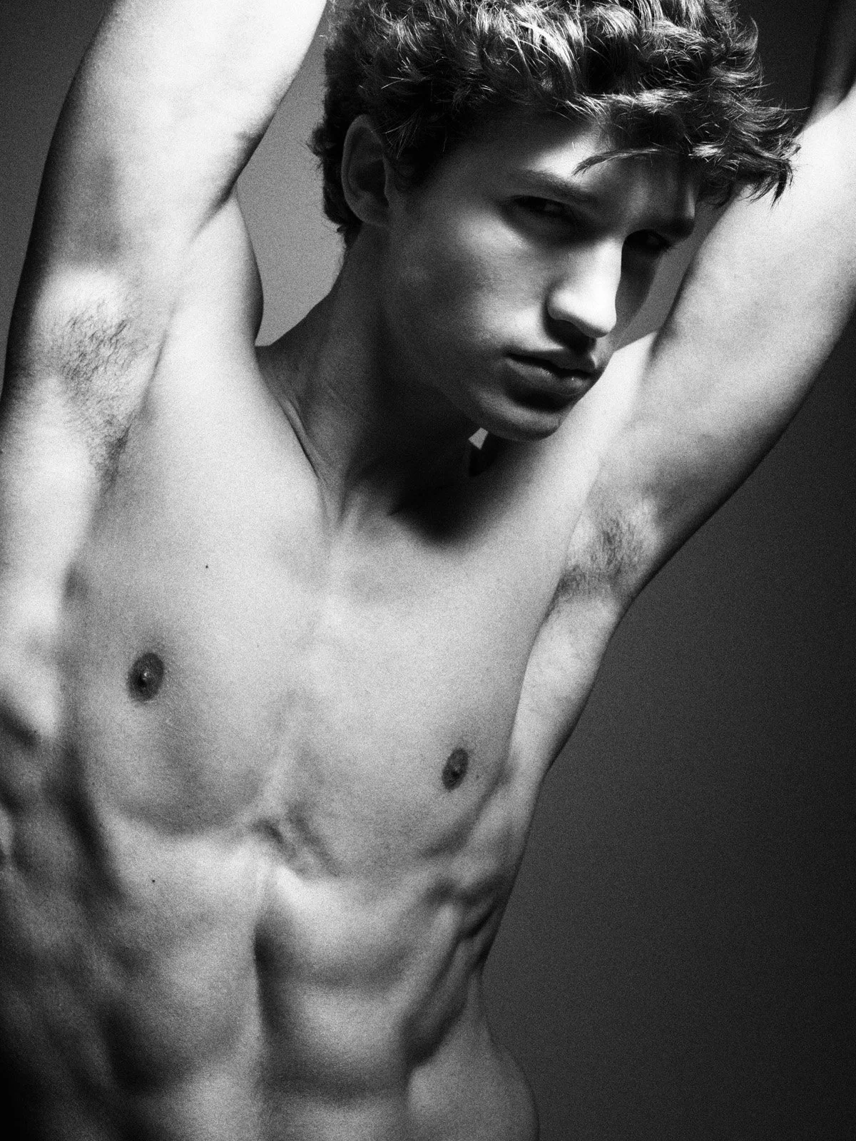 Black and white photo of a shirtless young man with tousled hair, posing with arms raised.