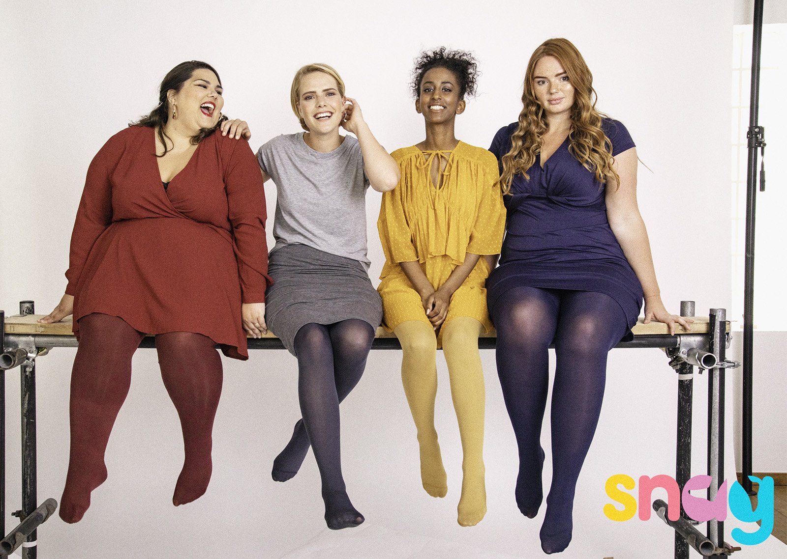Four diverse women seated on a bench, laughing and smiling, in front of a white background with colorful text. They are wearing colorful outfits and tights.