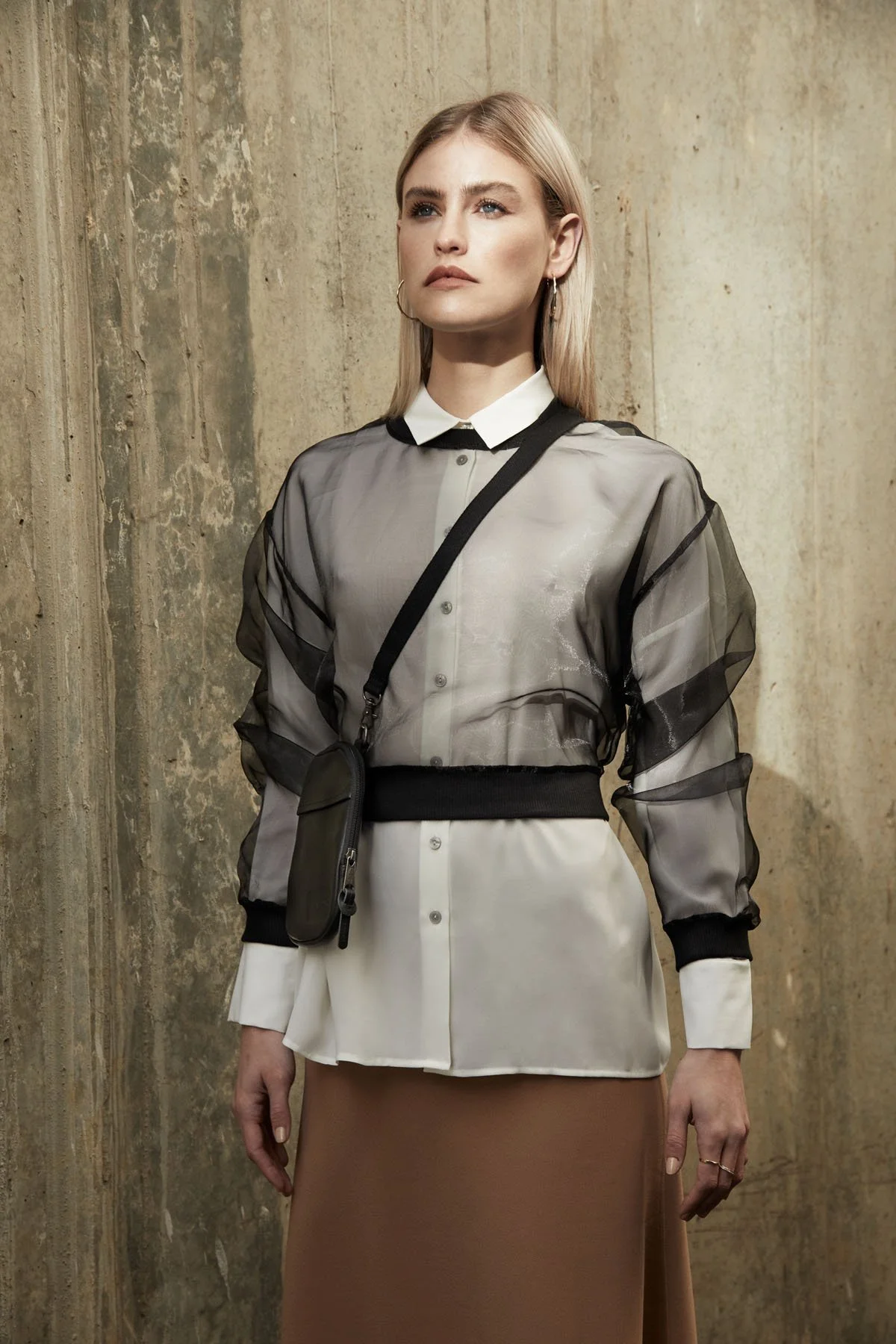 A woman with blonde hair and hoop earrings standing against a textured concrete wall. She is wearing a sheer black and white button-up top over a white shirt, a beige skirt, and has a small black crossbody bag.