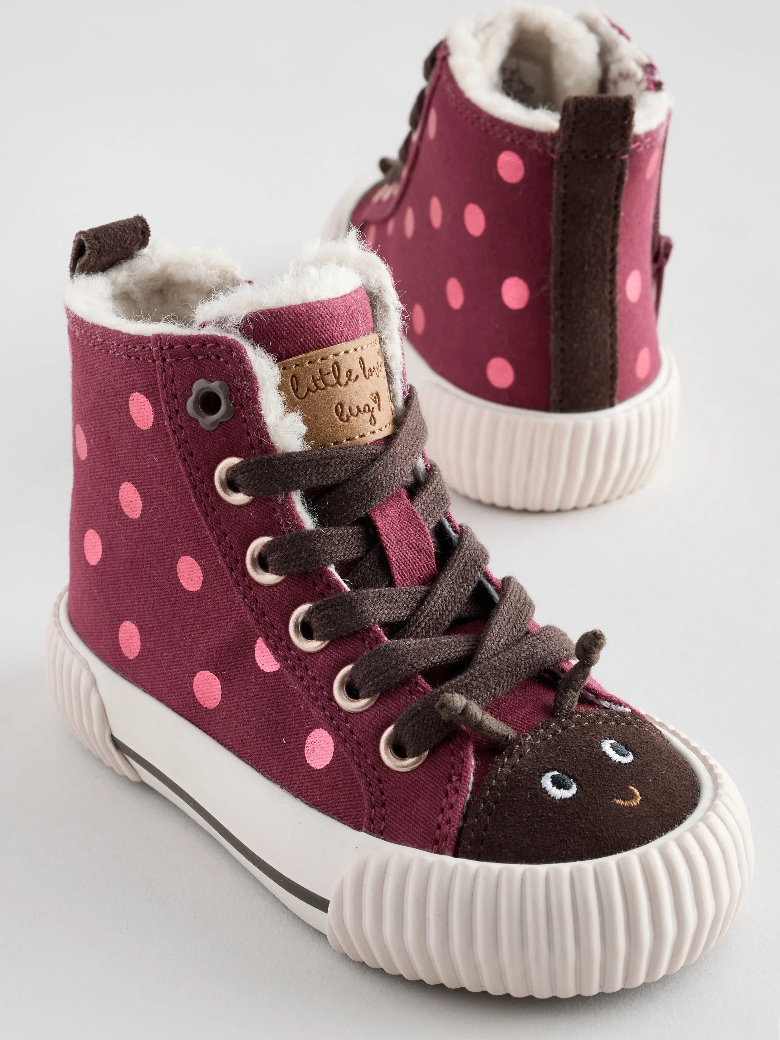 Pink children's high-top sneaker with polka dots, brown laces, and a smiling face design on the toe in white, black, and peach, featuring plush lining and a textured white sole.