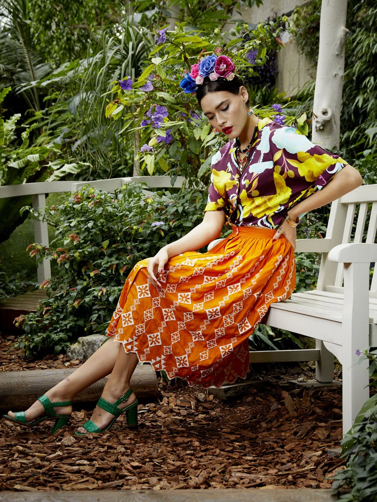 Woman sitting on a white garden bench in a lush, green garden, wearing a colorful floral shirt, orange patterned skirt, green high-heeled sandals, and a floral headpiece.