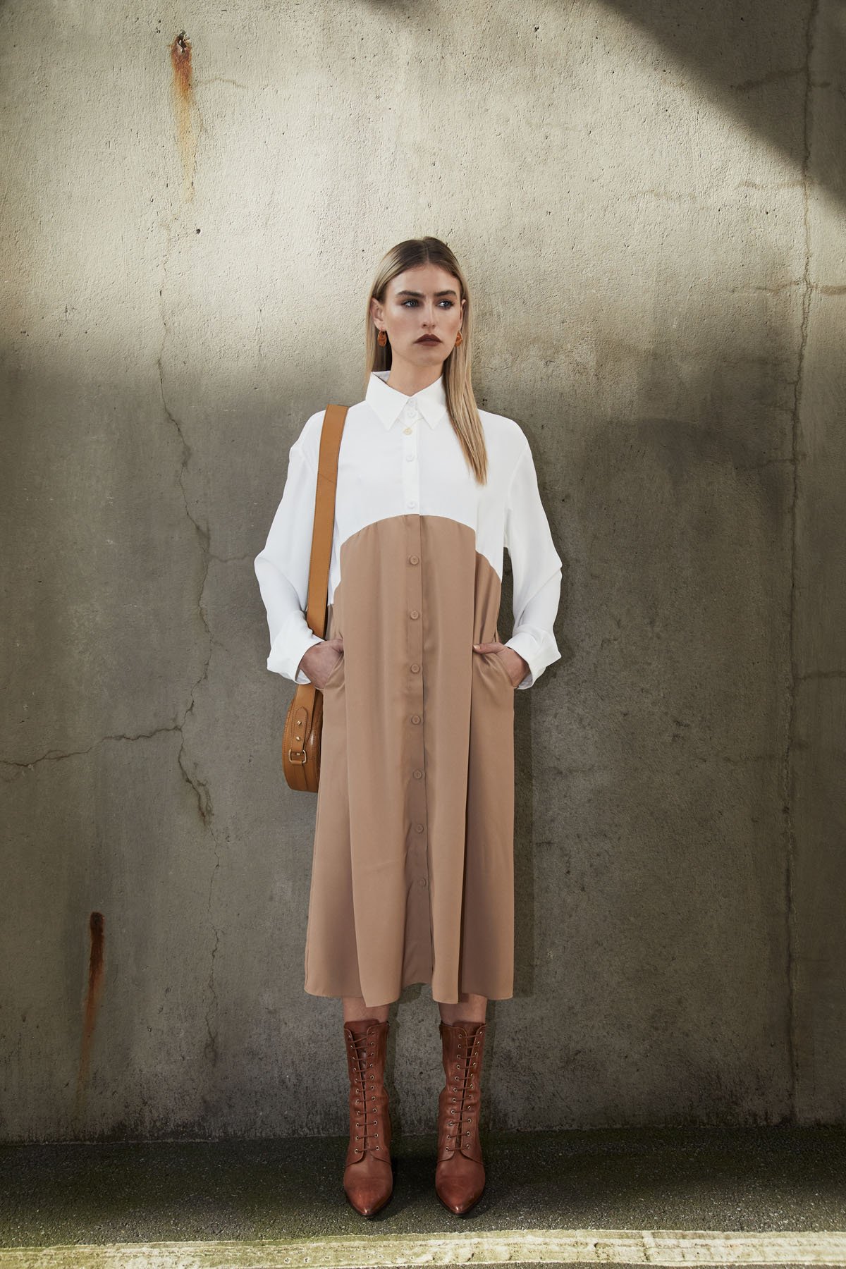 A woman standing against a concrete wall with cracks, wearing a white blouse with a brown apron over it, brown lace-up boots, and carrying a brown shoulder bag.
