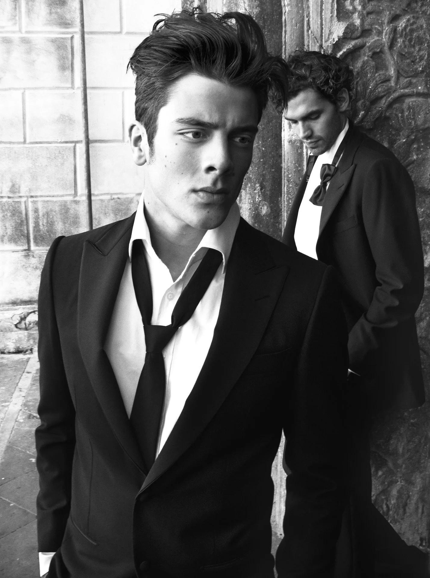 Two young men dressed in suits and ties, standing against a brick wall, one facing forward with a serious expression, the other looking downward with closed eyes.
