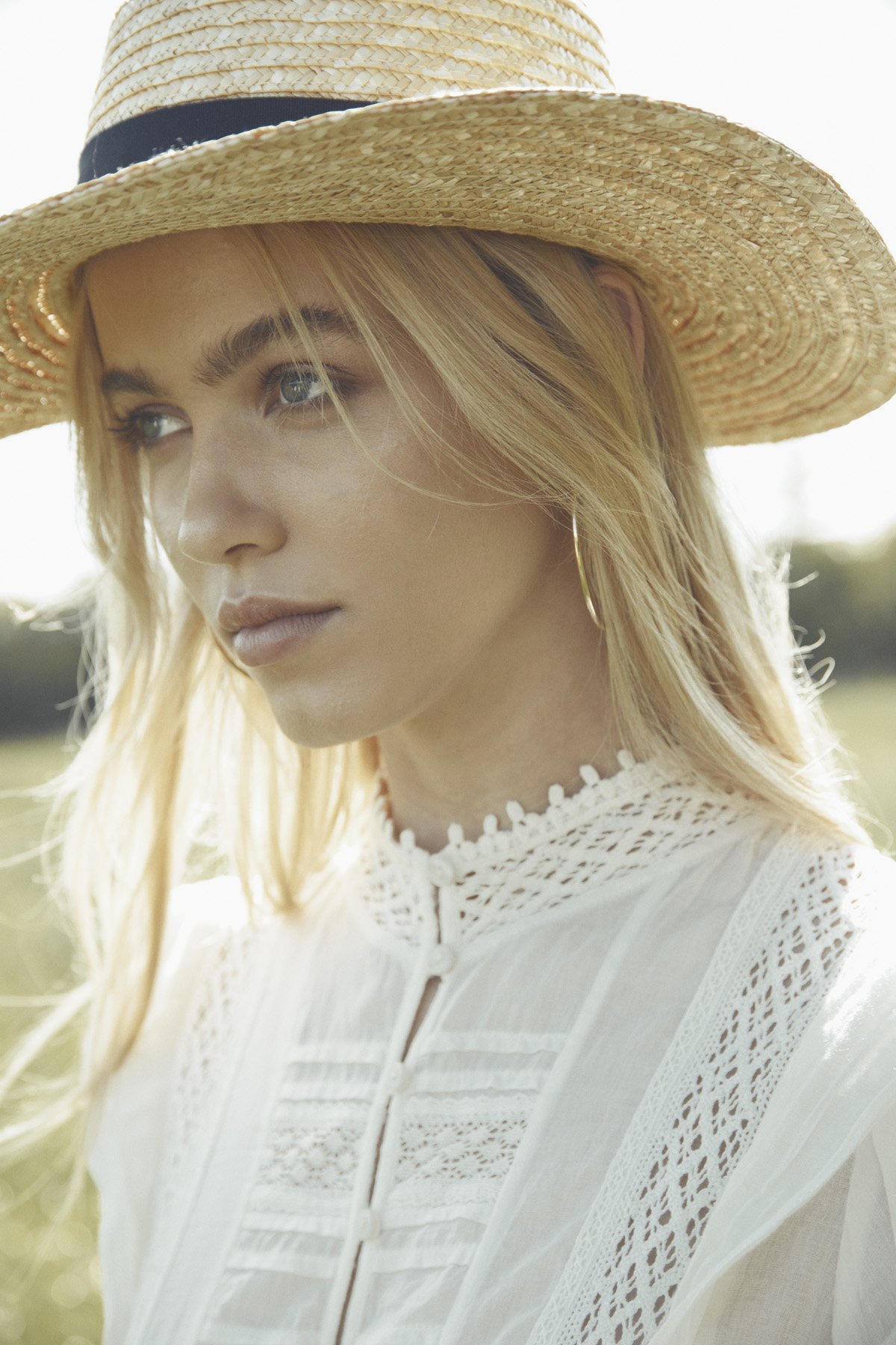 A young woman with blonde hair and blue eyes, wearing a large straw sun hat and a white lace blouse.