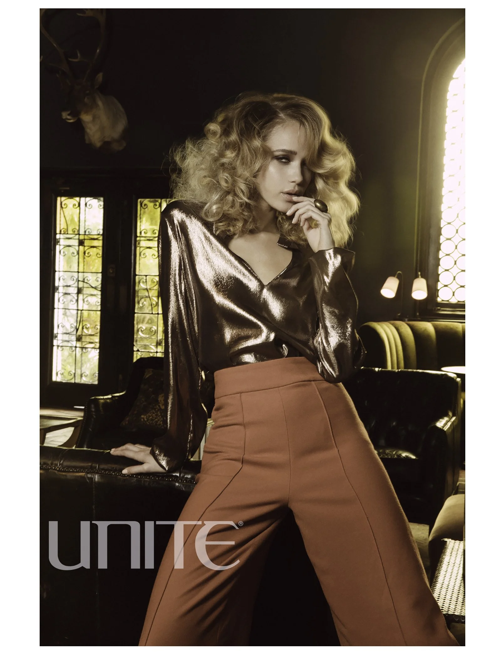 A woman with curly blonde hair in a shiny bronze blouse and high-waisted rust-colored pants, standing in a dimly lit room with stained glass windows, a black leather couch, and a mounted deer head on the wall.