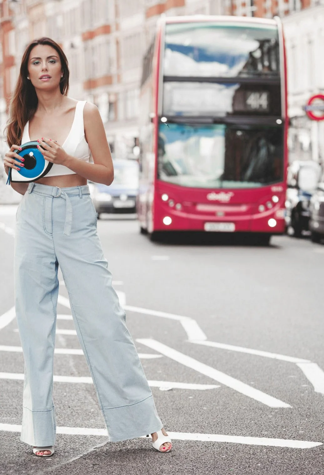 A woman standing in the street holding a circular object, with a pink double-decker bus and other vehicles in the background.
