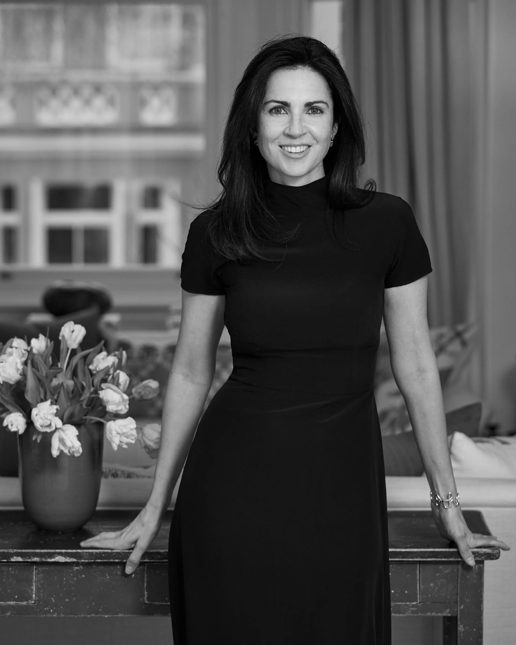 Black and white photo of a woman sitting on a wooden table. She has long dark hair and is wearing a black dress with a high neckline. She is smiling and looking directly at the camera. Flowers in a vase and books can be seen in the background, along 