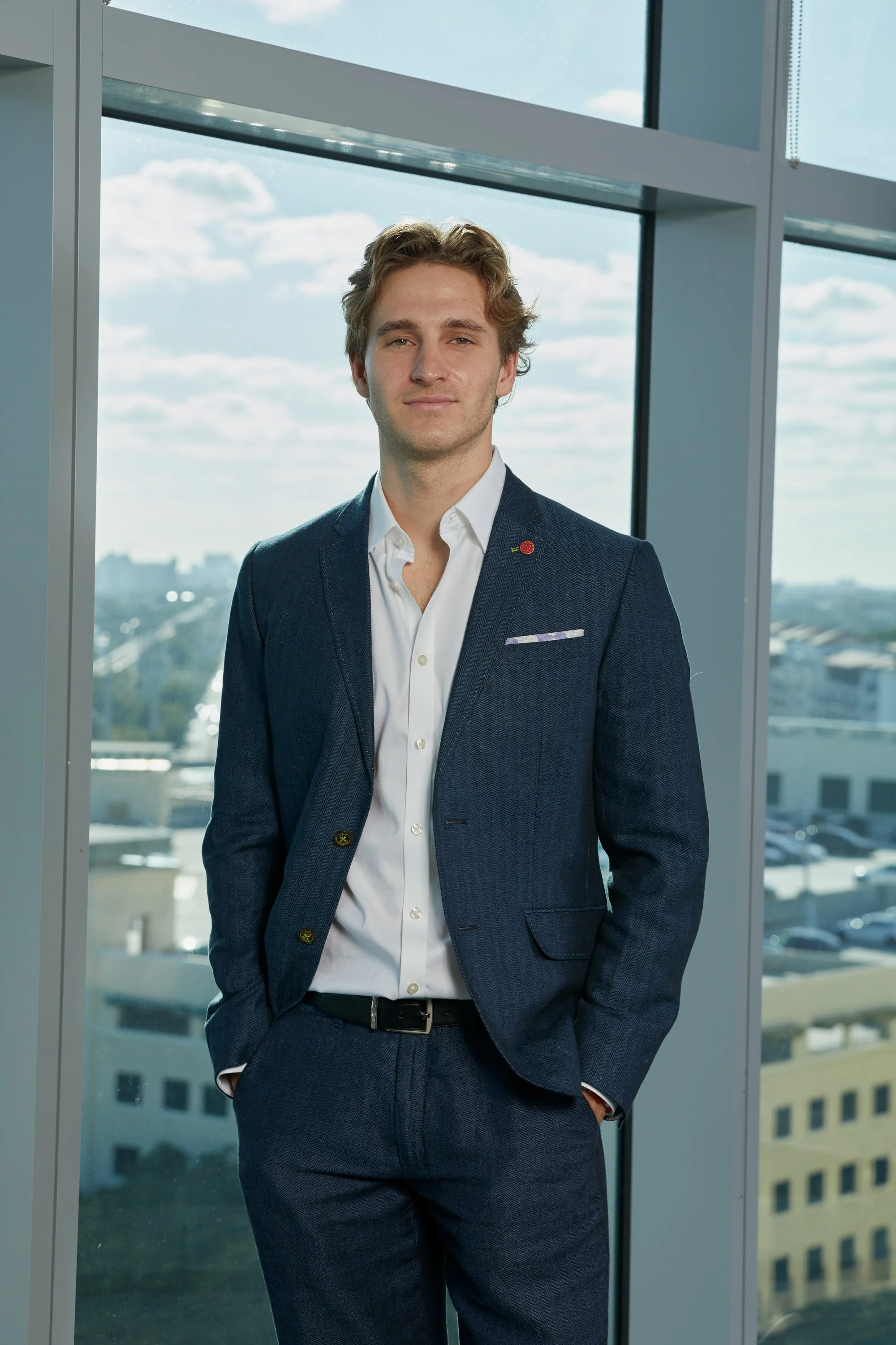A young man in a dark blue suit and white dress shirt standing in front of large windows in a modern building, with a cityscape and cloudy sky visible outside.