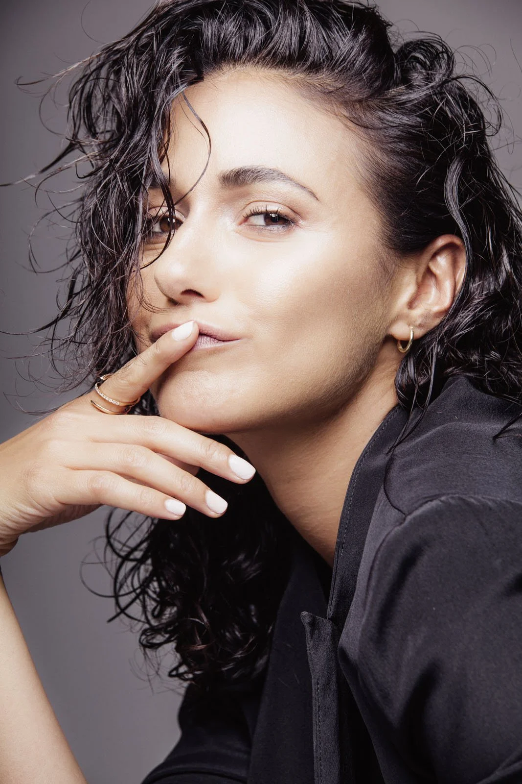 Close-up portrait of a woman with curly dark hair, light makeup, and piercings, touching her lips and looking at the camera.