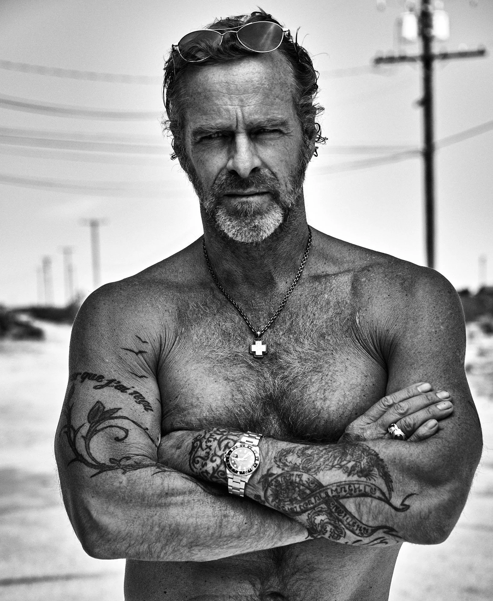 A shirtless, tattooed man with a beard, wearing sunglasses on his head, a necklace with a cross pendant, a wristwatch, and rings on his fingers, standing with crossed arms outdoors, possibly at the beach, in black and white.