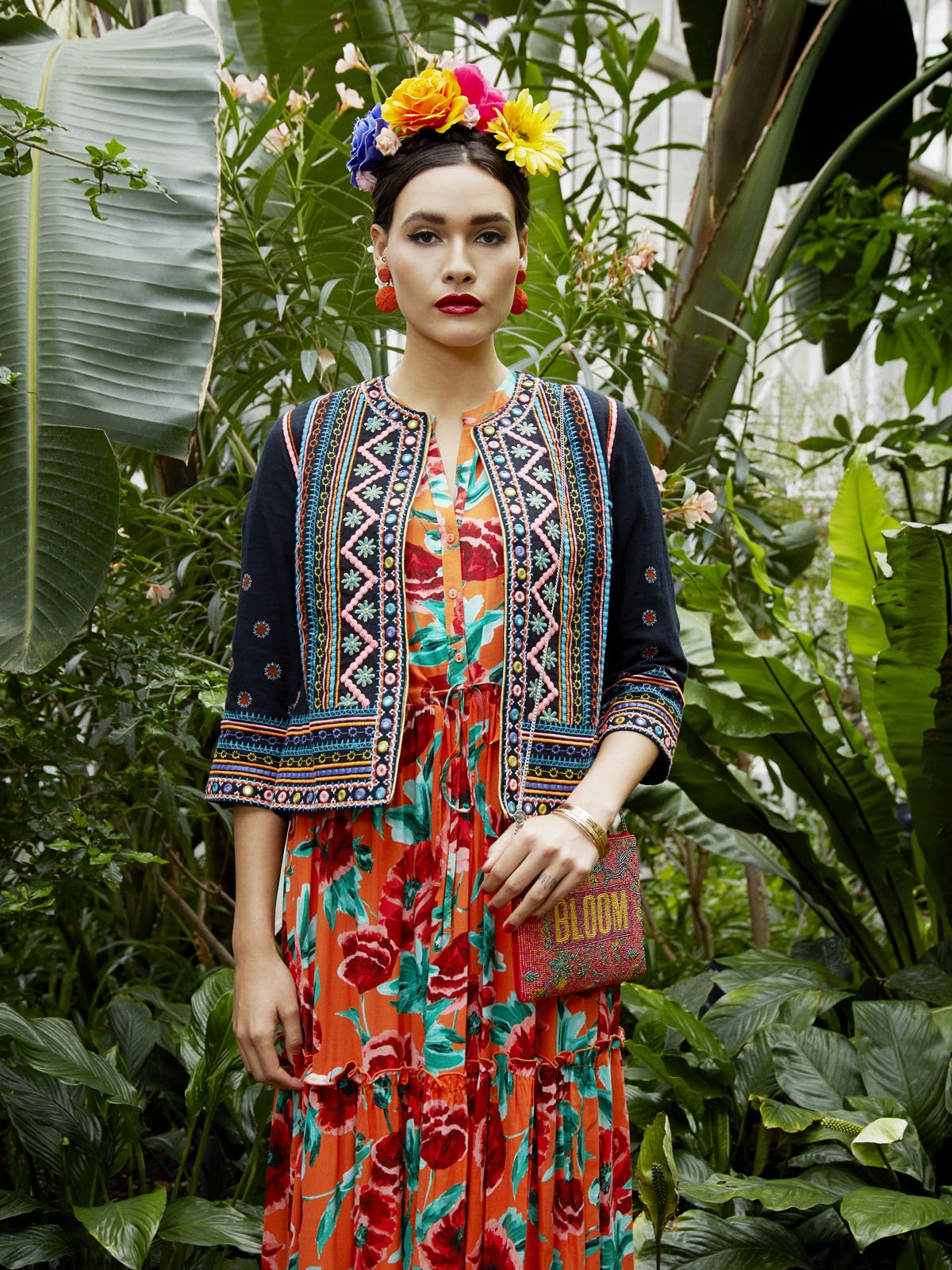 A woman with dark hair wearing a colorful floral dress, a navy embroidered jacket, and a flower crown, standing among green tropical plants.