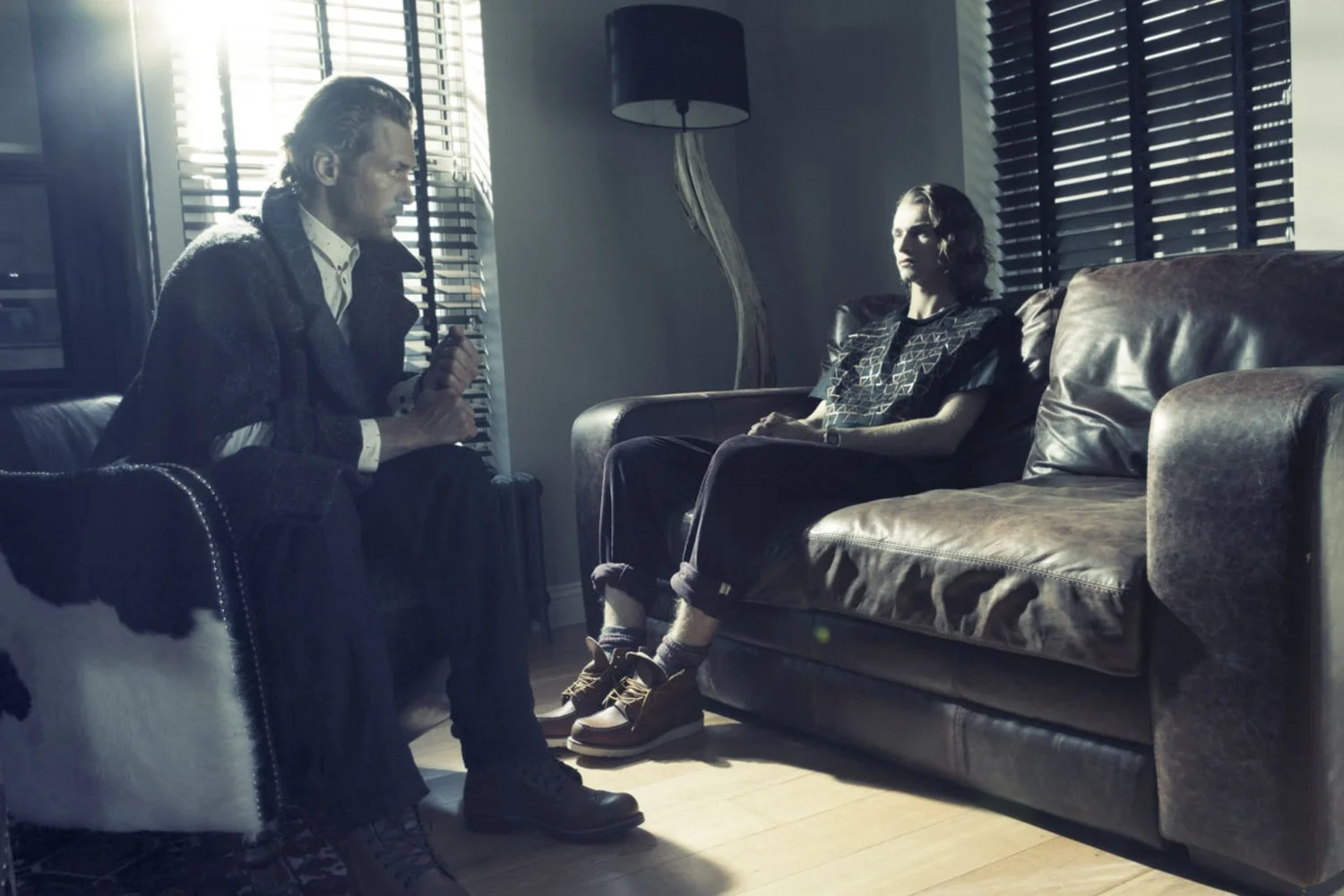 A man and woman sit in a dimly lit room with blinds. The man is seated on a chair on the left, leaning forward slightly, and the woman is seated on a brown leather couch on the right, with her hands folded in her lap.