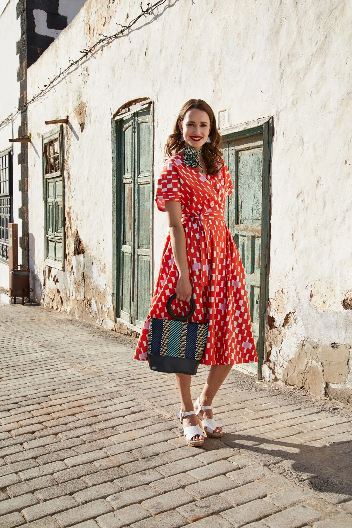 A woman in a red and white patterned dress standing on a cobblestone street, smiling, holding a woven handbag, in front of an old white wall with green wooden shutters on windows.