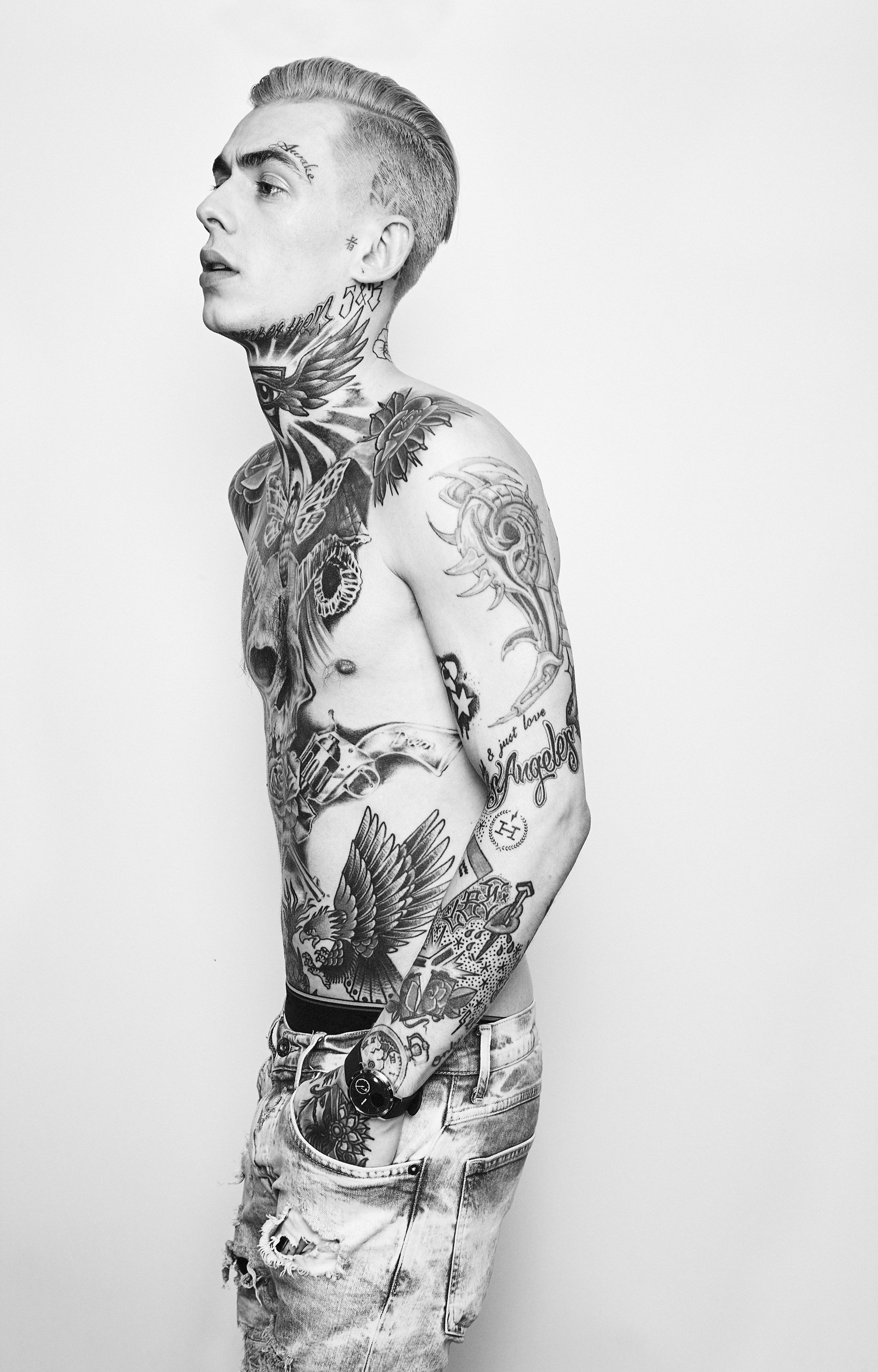 Black and white photo of a tattooed young man wearing jeans and a wristwatch, standing sideways against a plain background.
