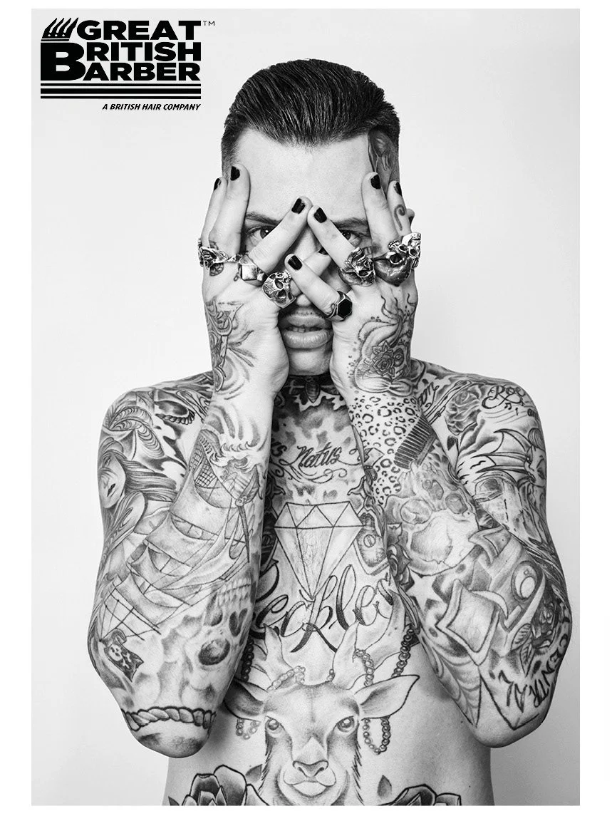 Black and white photo of a tattooed man covering his face with his hands, wearing multiple rings, with tattoos of various designs including a deer, a diamond, a ship, and words on his chest and arms. His hair is slicked back. Logo in the top left cor