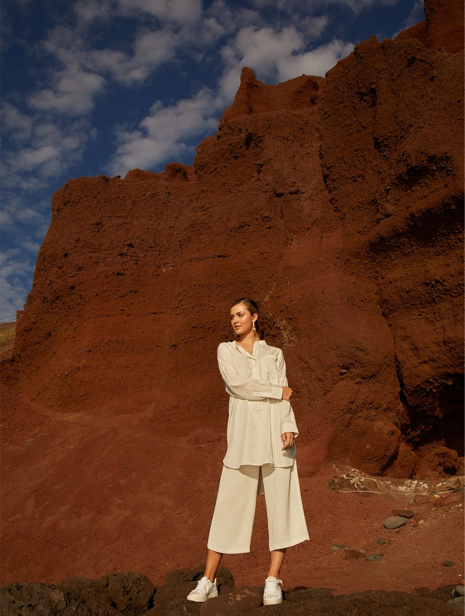 A woman in white clothing, including a blouse and wide-leg pants, stands with her arms crossed in front of large red rock formations under a blue sky with scattered clouds.