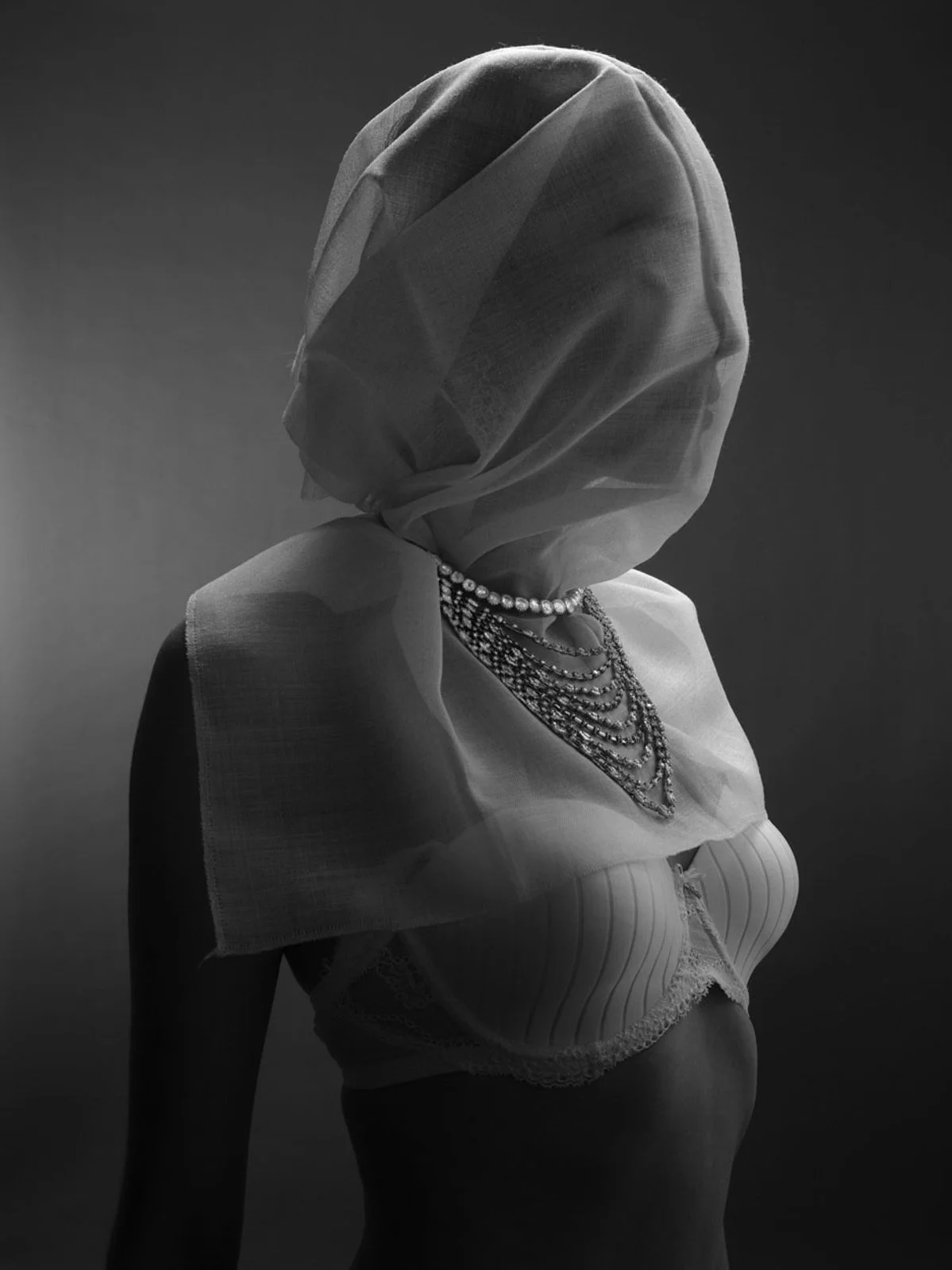 Black and white photo of a woman wearing layered pearl and gemstone necklaces, a striped bustier, and a sheer headscarf covering her face and head, with a plain dark background.