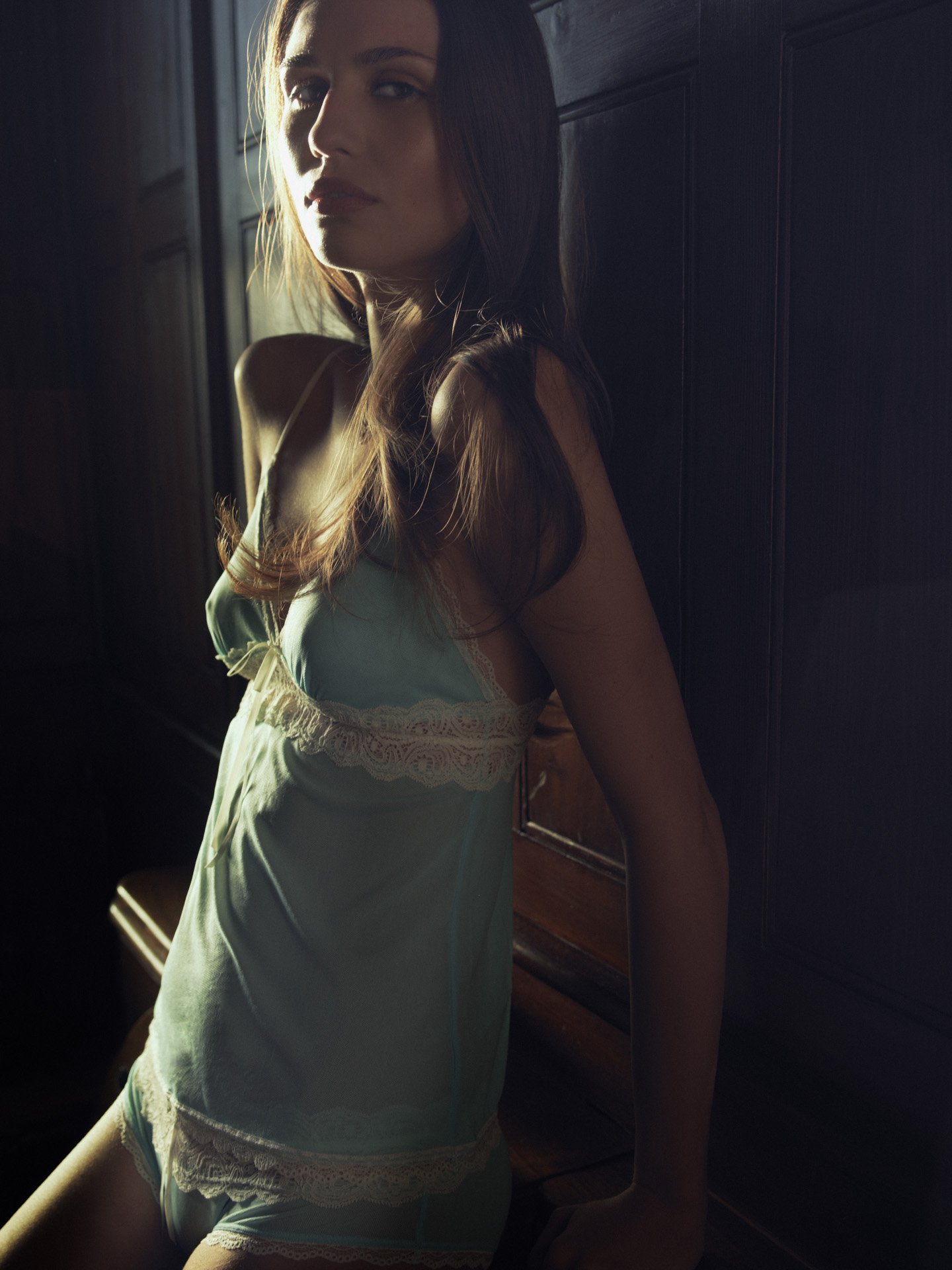 A woman with long brown hair sitting in a dimly lit room, wearing a light blue nightgown with lace trim, looking at the camera with a serious expression.