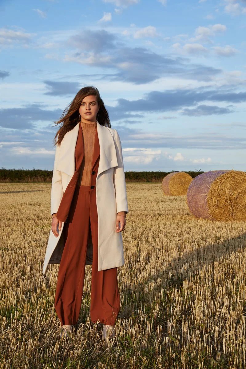A woman standing in a harvested wheat field with hay bales and a partly cloudy sky in the background.