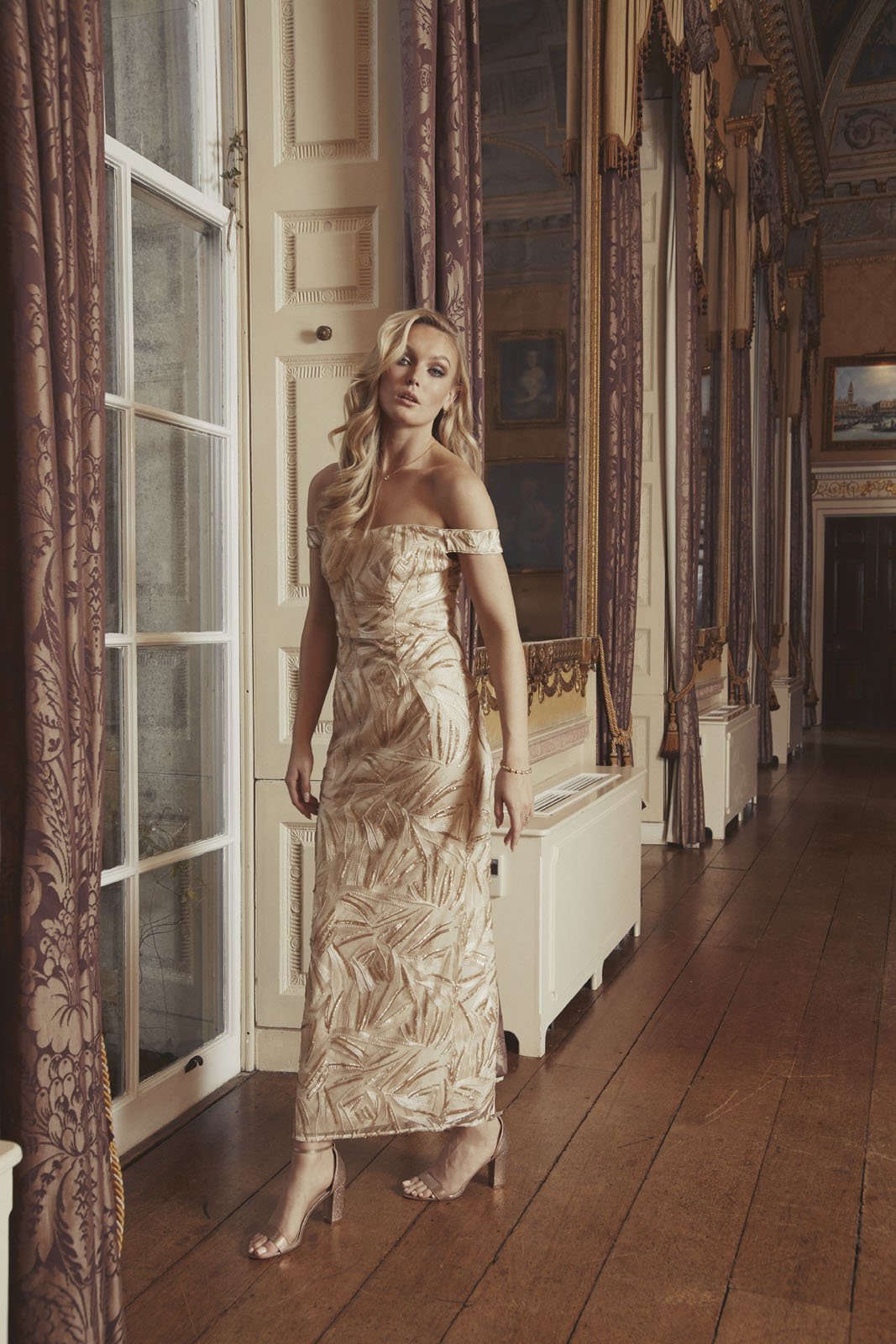 A woman in a gold patterned off-shoulder dress and high heels standing in an ornate room with tall windows, draped curtains, and gilded decor.