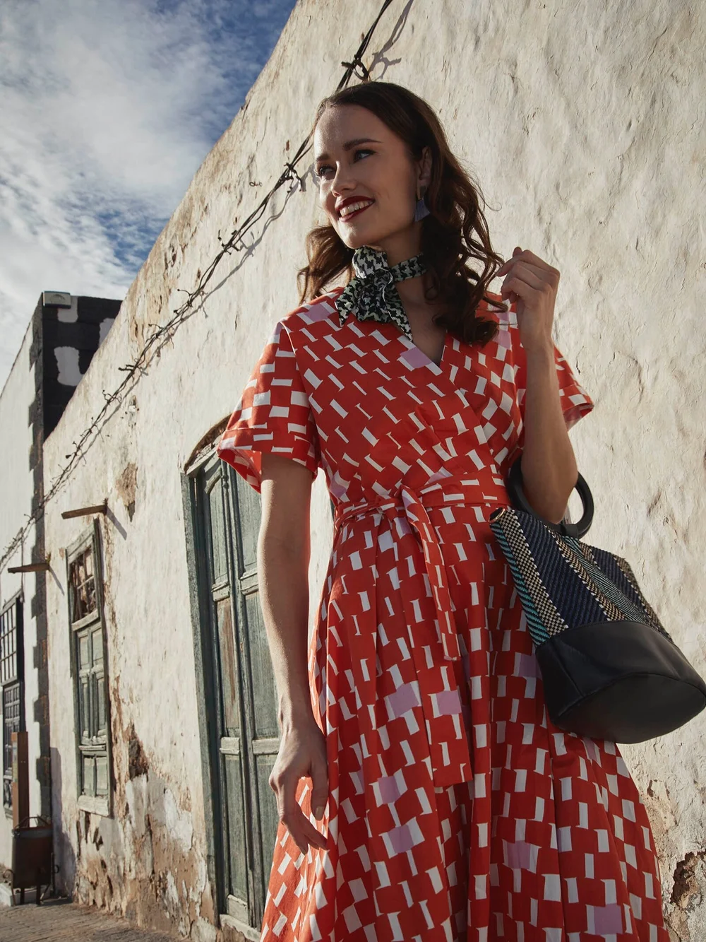 A woman with dark wavy hair wearing a red and white patterned dress, a black and white scarf, and earrings, smiling while standing near a white wall with windows and a barbed wire on top against a blue sky.